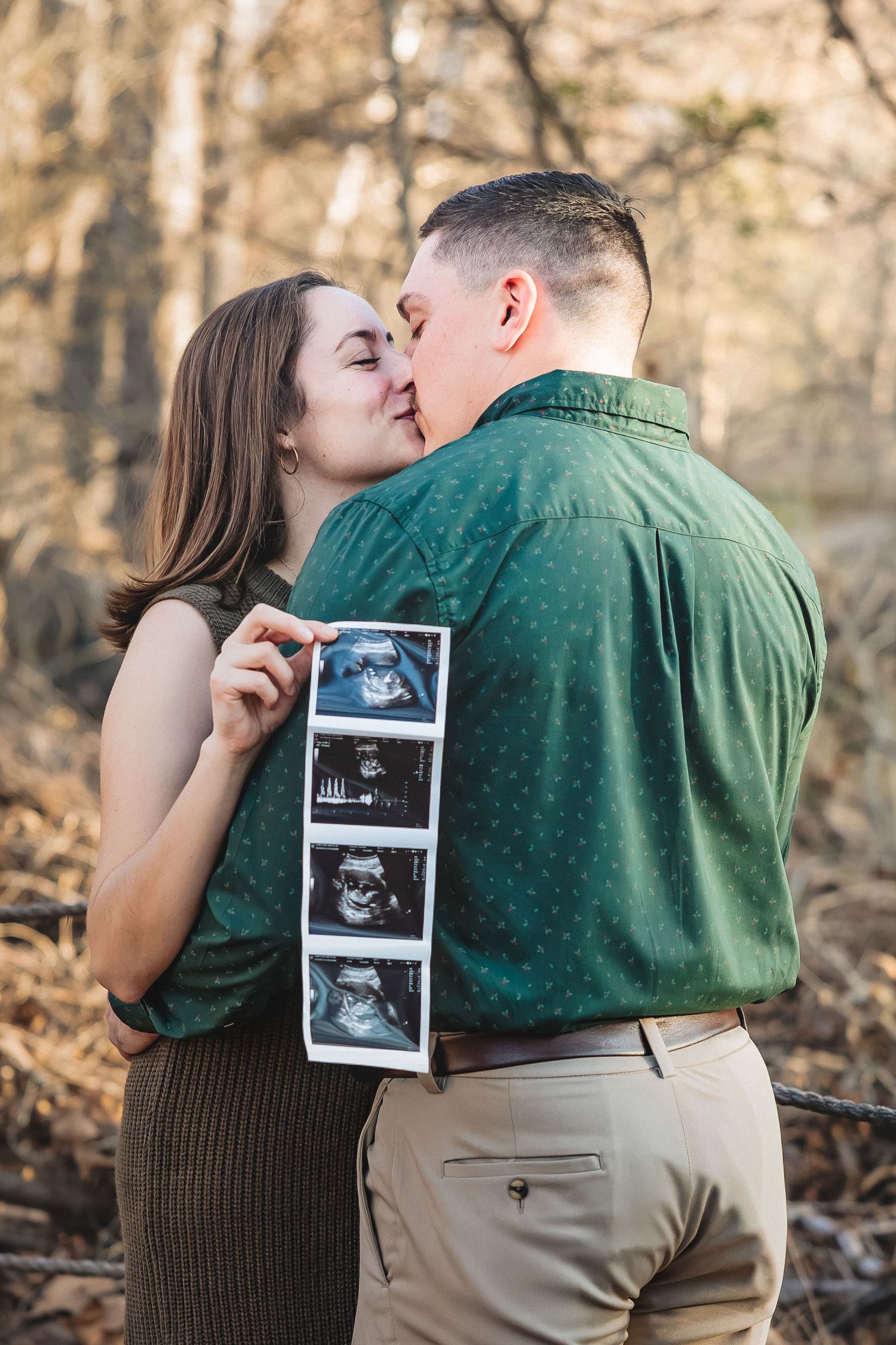 A couple shares a tender kiss in the woods while holding ultrasound photos during their pregnancy announcement photoshoot.