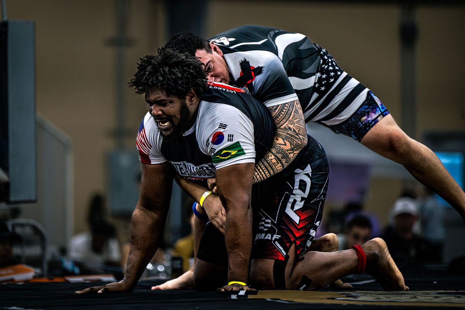 Two athletes compete in a grappling match at an indoor martial arts tournament, wearing team jerseys.
