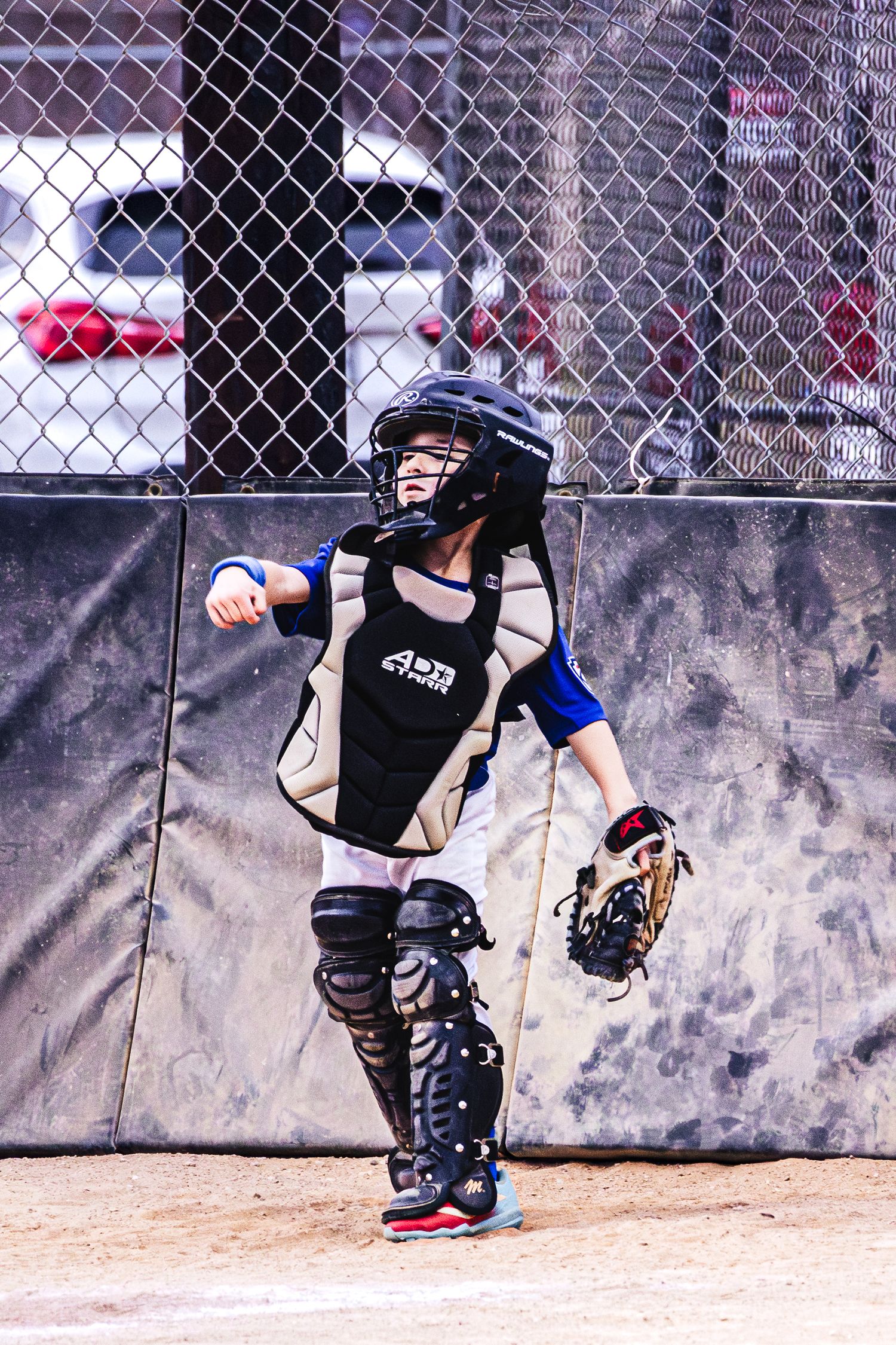 Young baseball catcher in full gear including helmet, chest protector, and shin guards, ready to play on a dirt field.