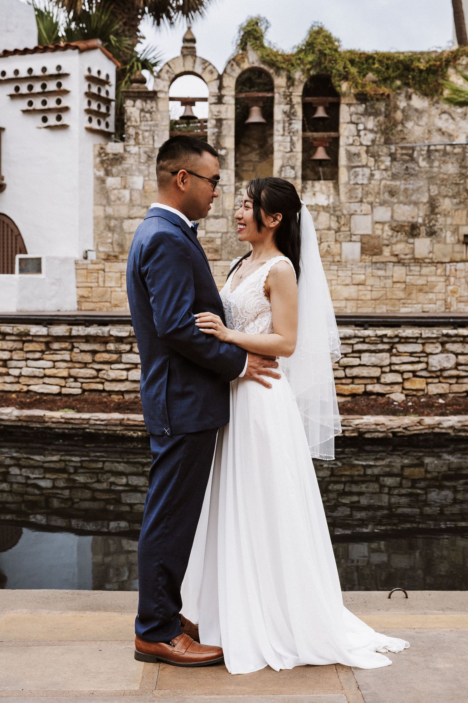 A romantic wedding couple sits on stone steps near a historic mission wall with white arches and bells.