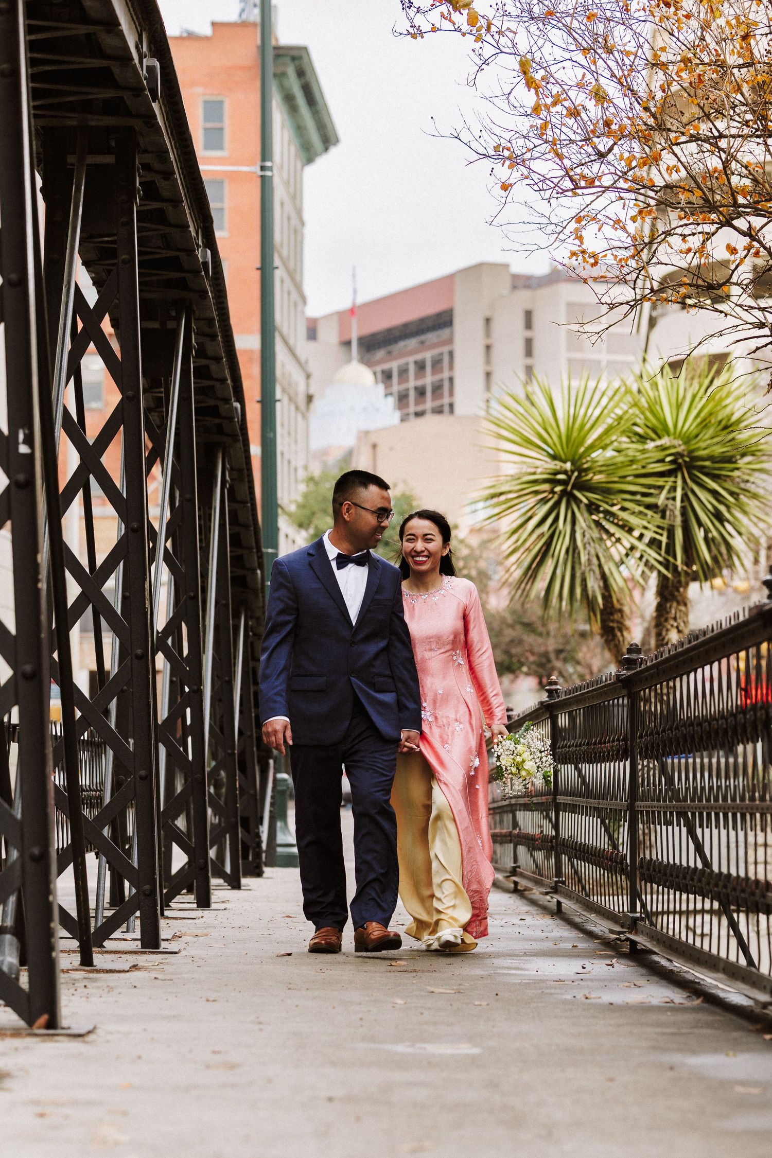 A couple walks along an iron-railed walkway with autumn trees and urban buildings in the background.