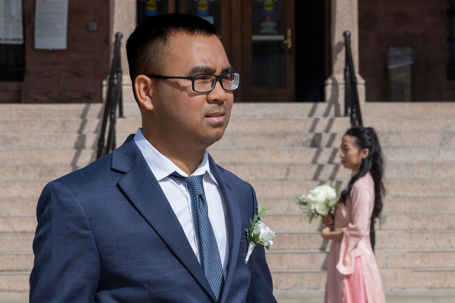 A wedding party poses on stone steps outside a brick building, with people wearing formal attire and floral boutonnieres.