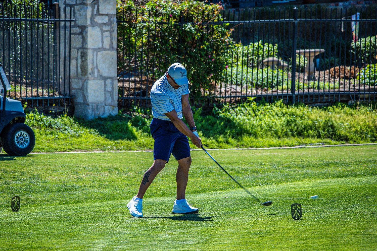 Golfer in blue shirt and navy shorts swinging a golf club on a sunny green course near a stone fence.