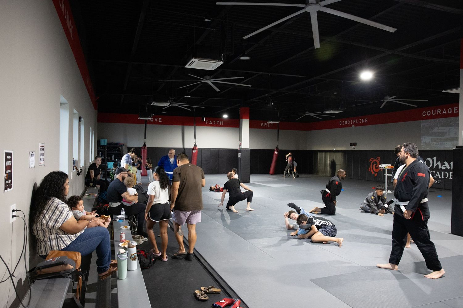 Students and spectators gather in a martial arts training gym during a class session on a large gray mat.