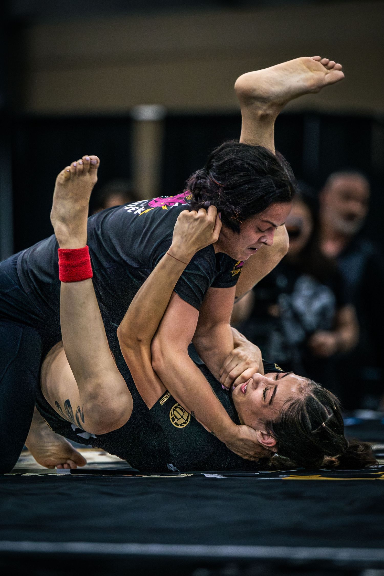Two women competing in a grappling martial arts match on a black mat during a BJJ or MMA competition.