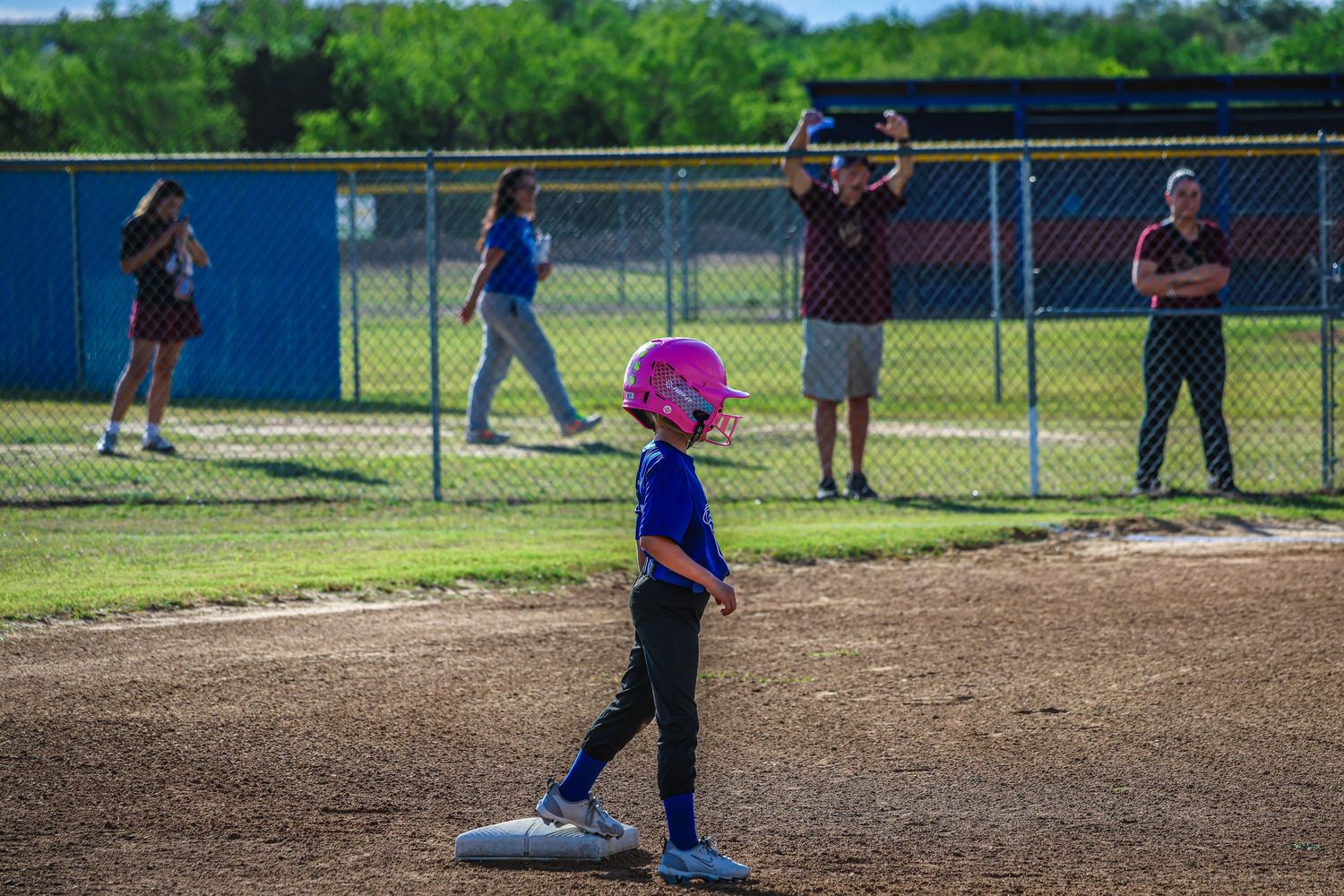 Young girl in pink helmet stands on base during youth softball game while coaches and parents watch from the sideline.