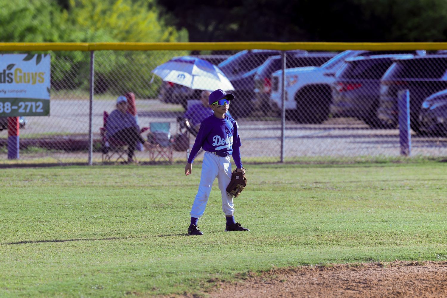 Youth baseball player in Dodgers uniform holding umbrella in outfield on sunny day.
