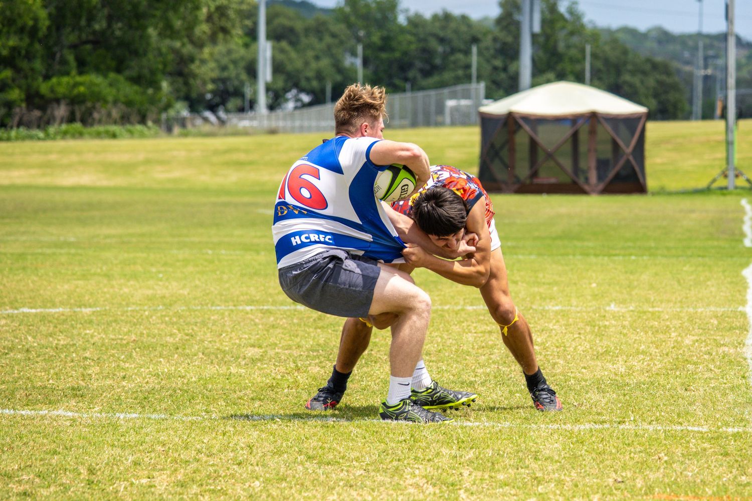 Two rugby players grapple during an outdoor match on a sunny green field.