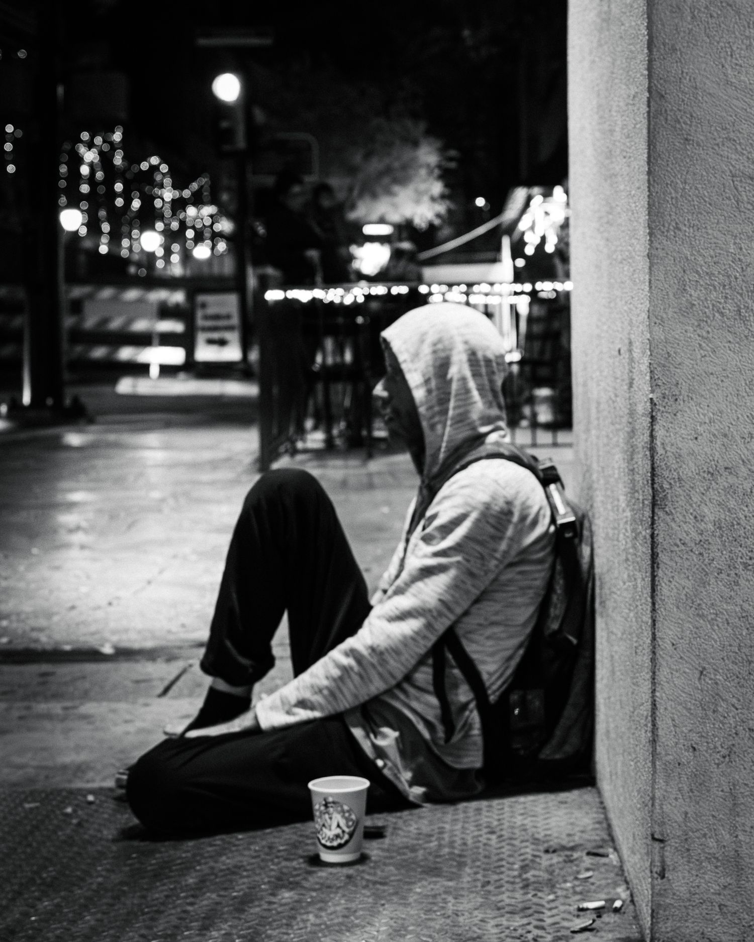 People sitting along urban sidewalk at night with city lights in background.