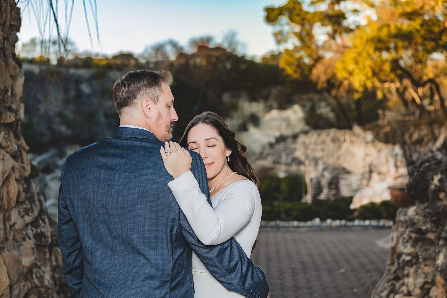 Newlyweds share intimate moment under rustic stone structure with fall foliage in background.