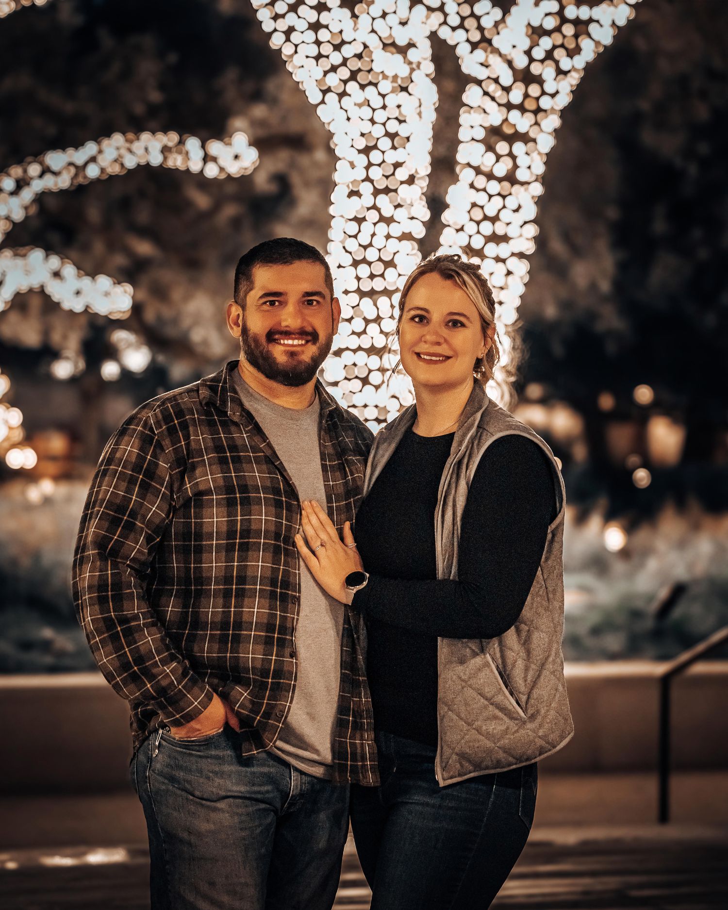 A couple stands together at night against a backdrop of twinkling bokeh lights in a romantic outdoor setting.
