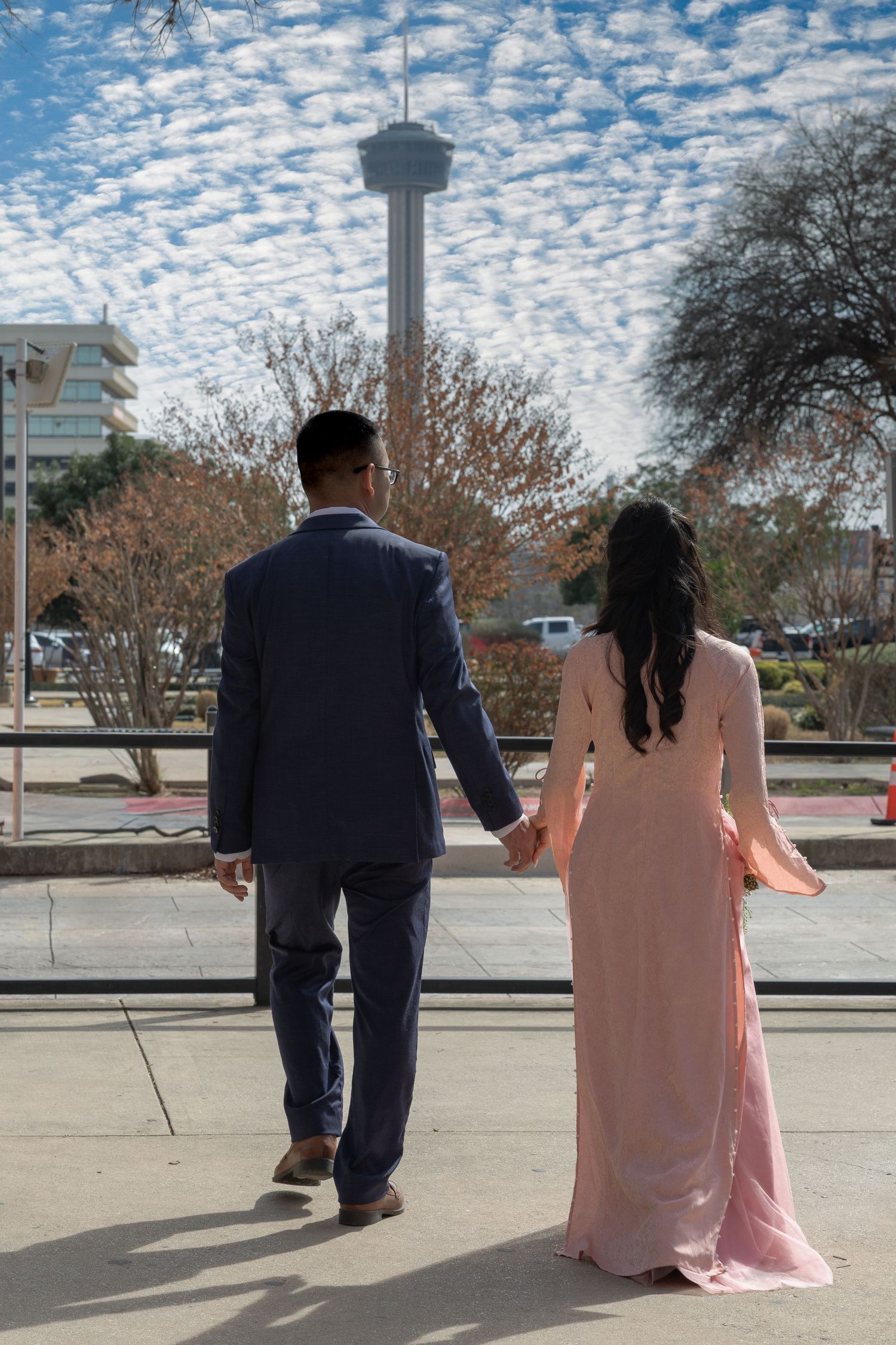 Couple holding hands walking toward Tower of the Americas in San Antonio, TX, woman in pink gown, man in navy suit.