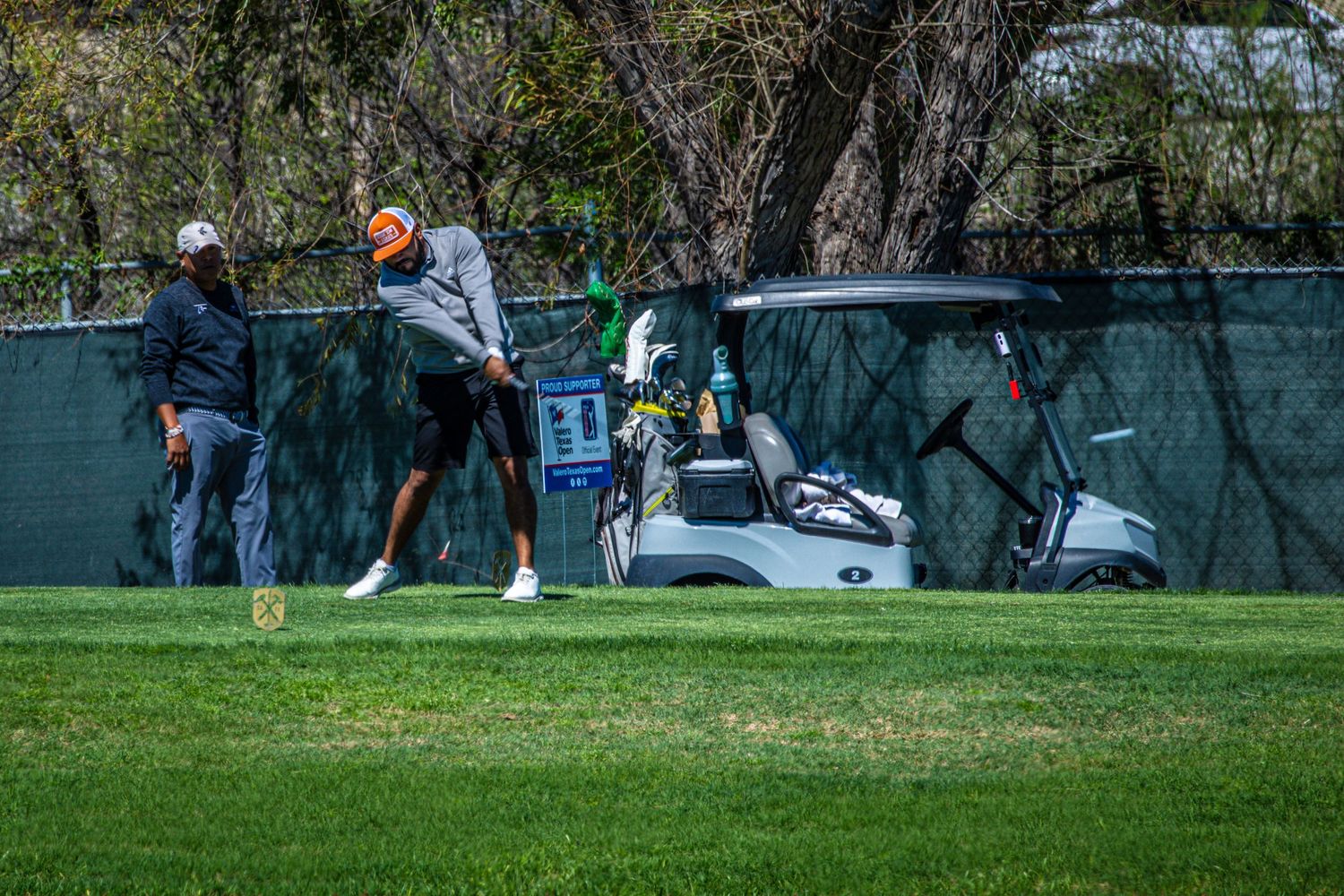 Two golfers on a lush green course near a golf cart, one mid-swing, with trees and a fence in the background.