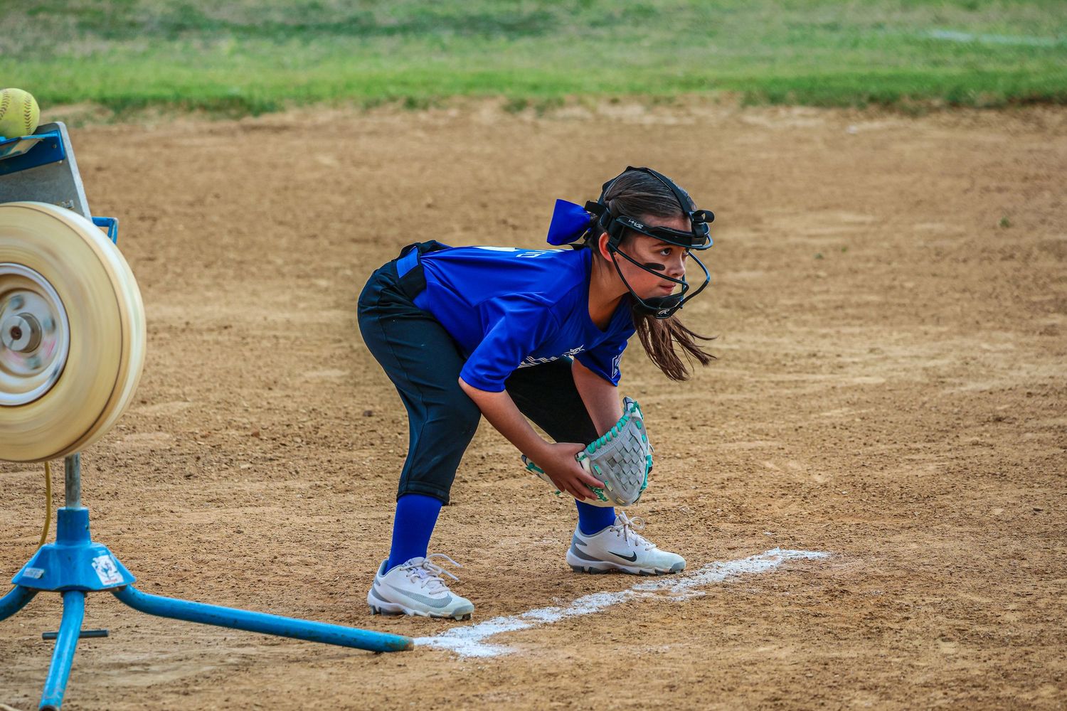 Female softball player in blue jersey crouches at first base, ready to field, with a pitching machine visible nearby.
