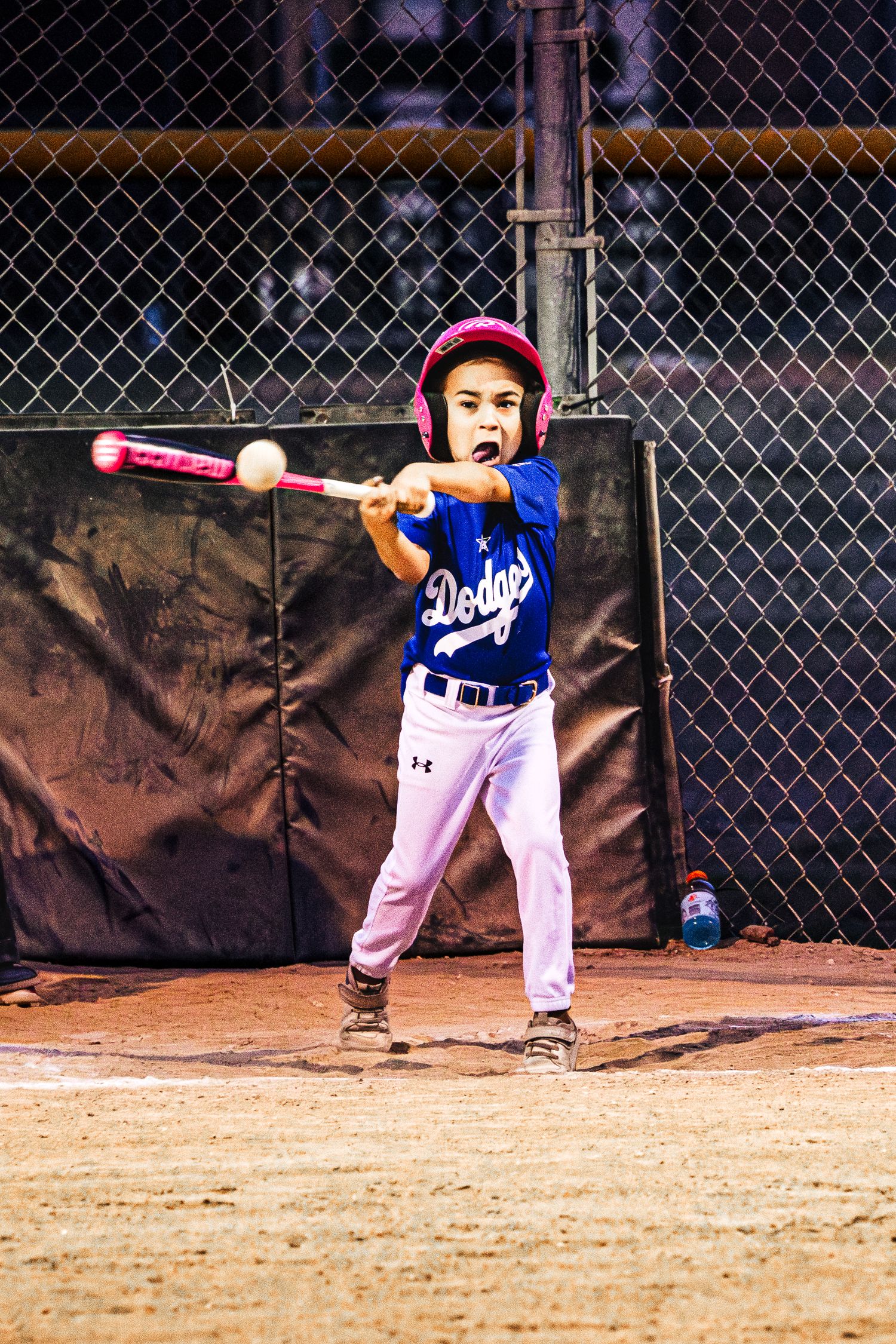 Young child in Dodgers uniform swinging a pink baseball bat during a little league night game.