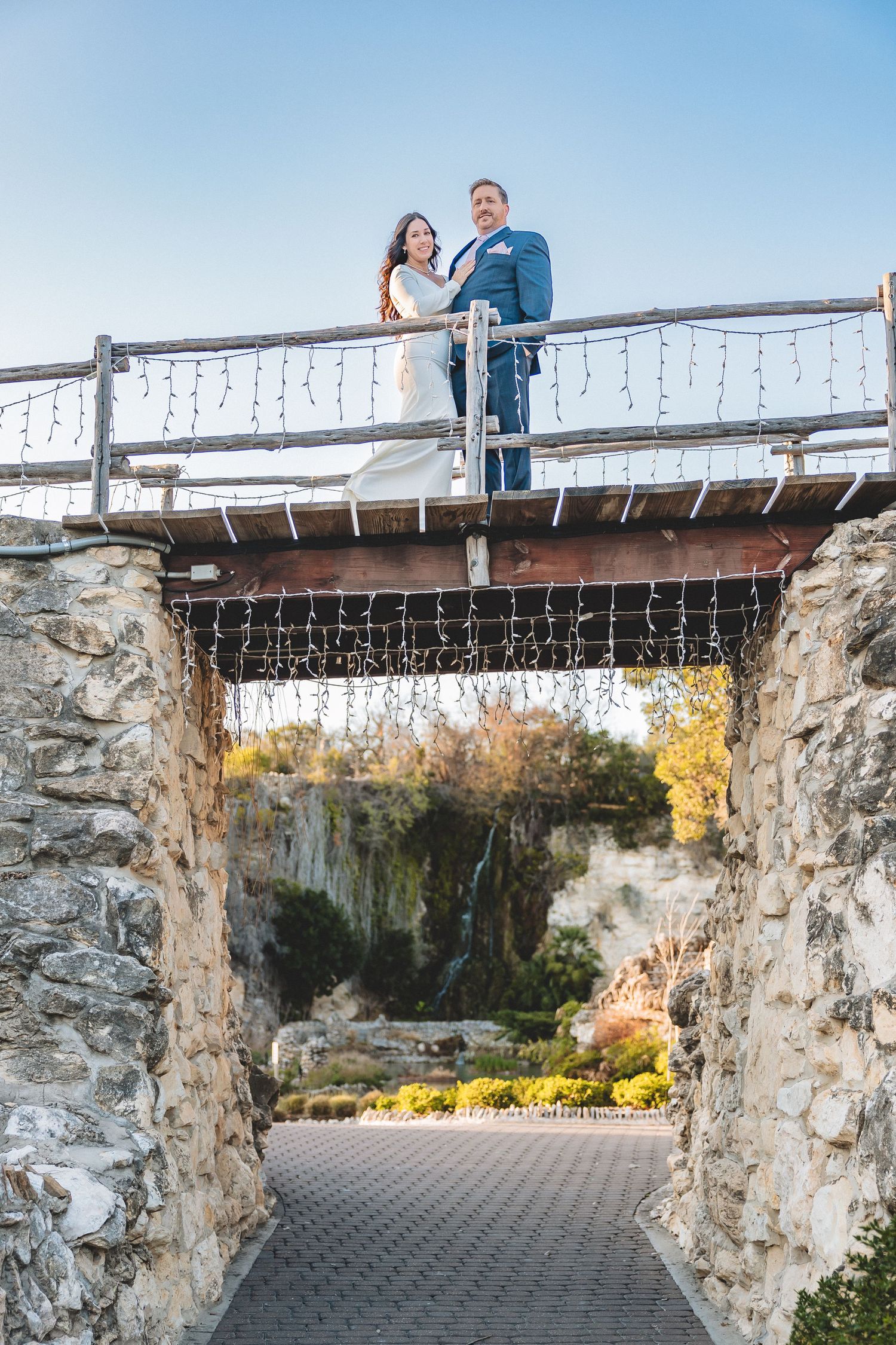 A couple stands on a rustic stone bridge with autumn foliage in the background during an outdoor photo session.