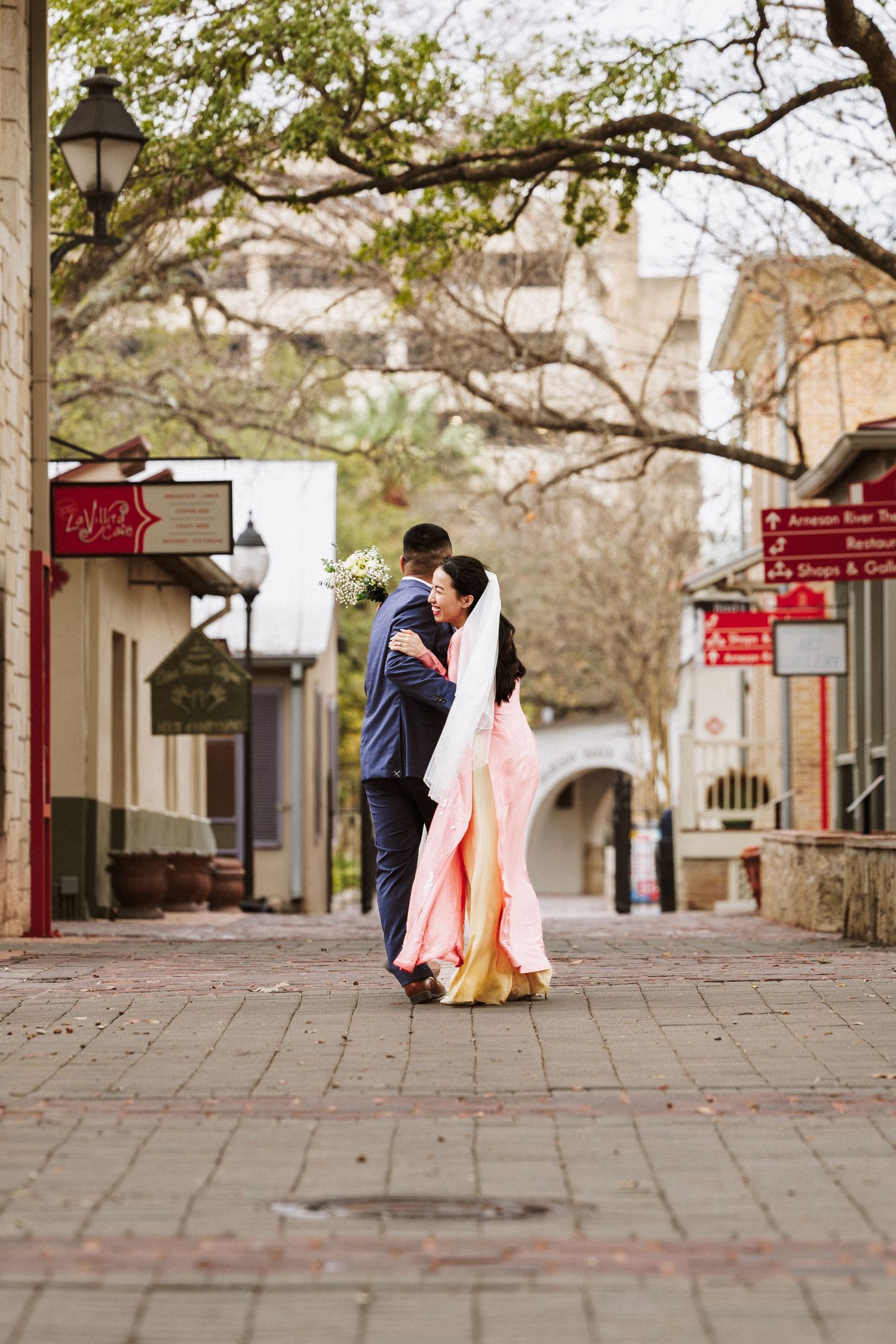 A couple embraces on a charming cobblestone street with historic architecture and lamp posts in the background.