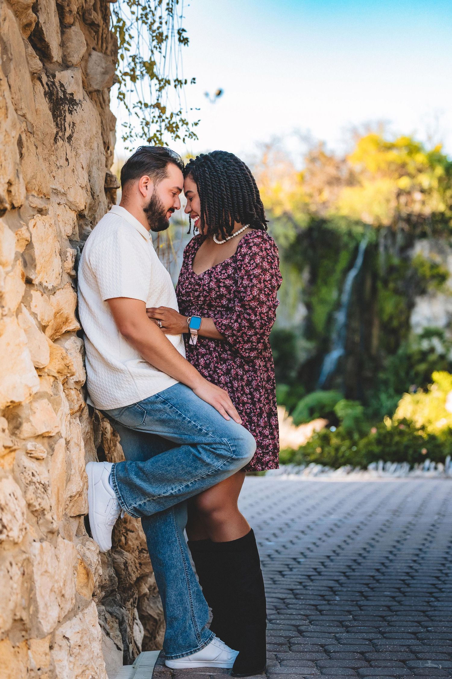 Romantic couple embraces against stone wall in a sunlit garden setting with lush greenery in background.