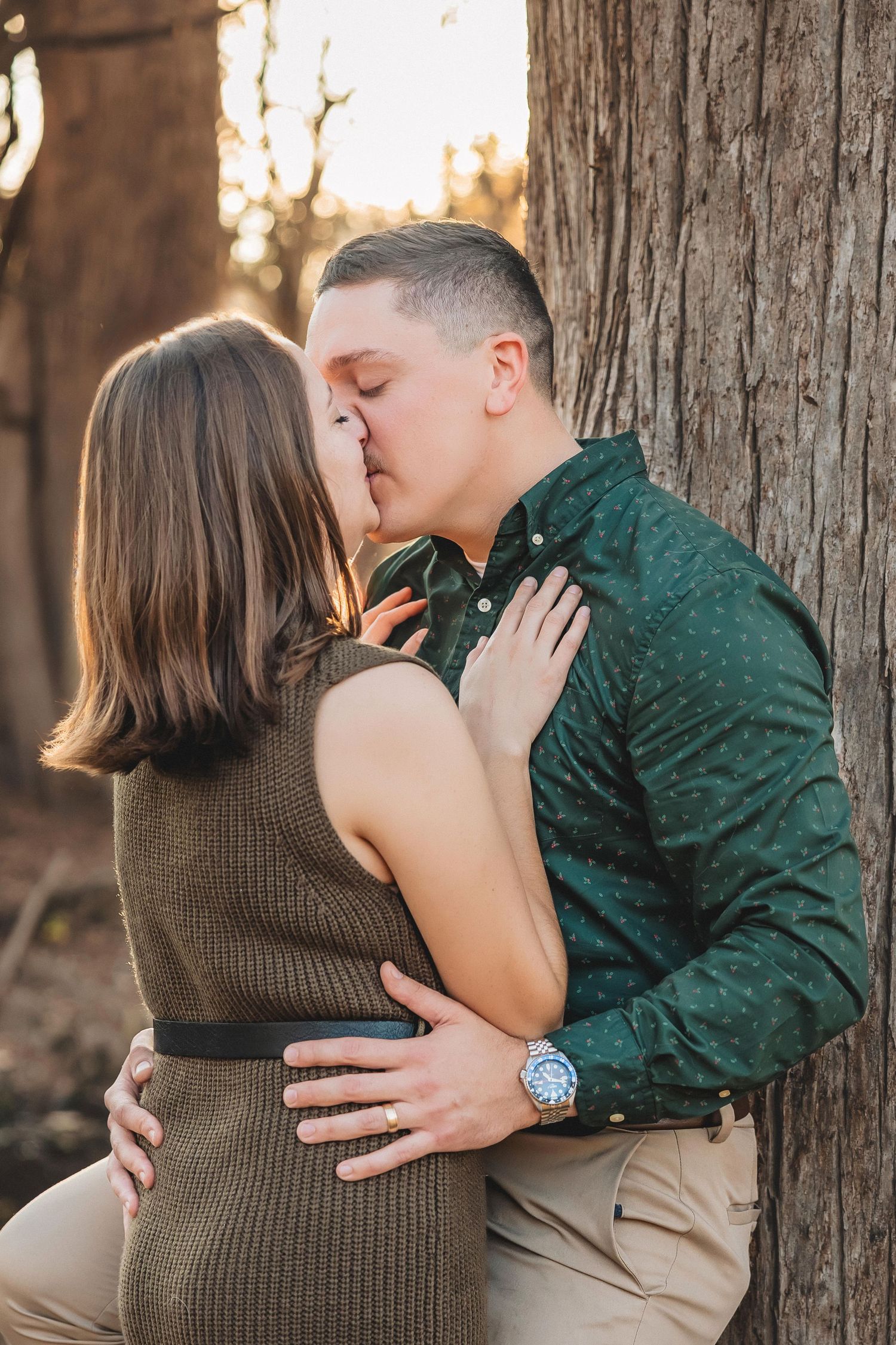 A romantic couple shares tender kisses in a sequence of four photos taken outdoors against rustic tree trunks at sunset.