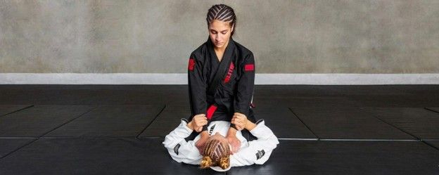 A BJJ practitioner in a black gi performs a ground technique on a white-gi opponent on black mats.
