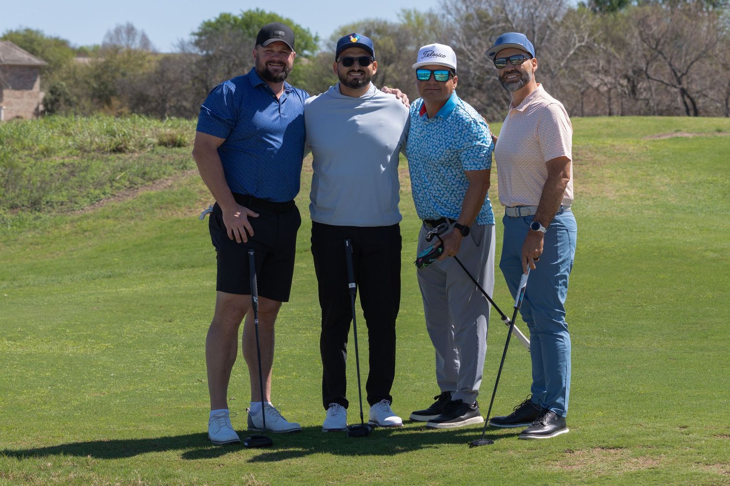 Four men holding golf clubs pose together on a sunny golf course with green fairways and blue sky.