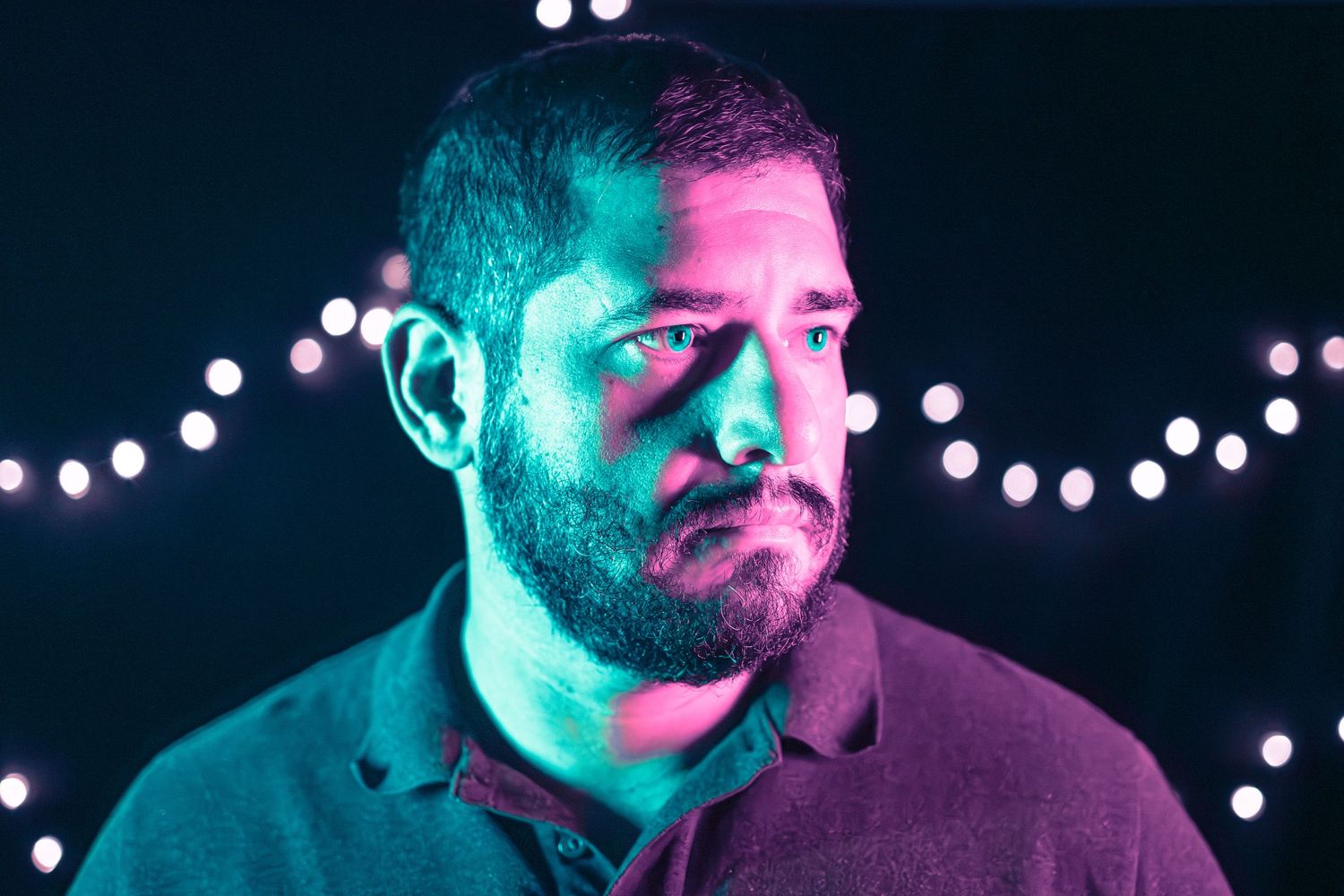 Man with beard under teal and pink neon lights with bokeh string lights in dark background.