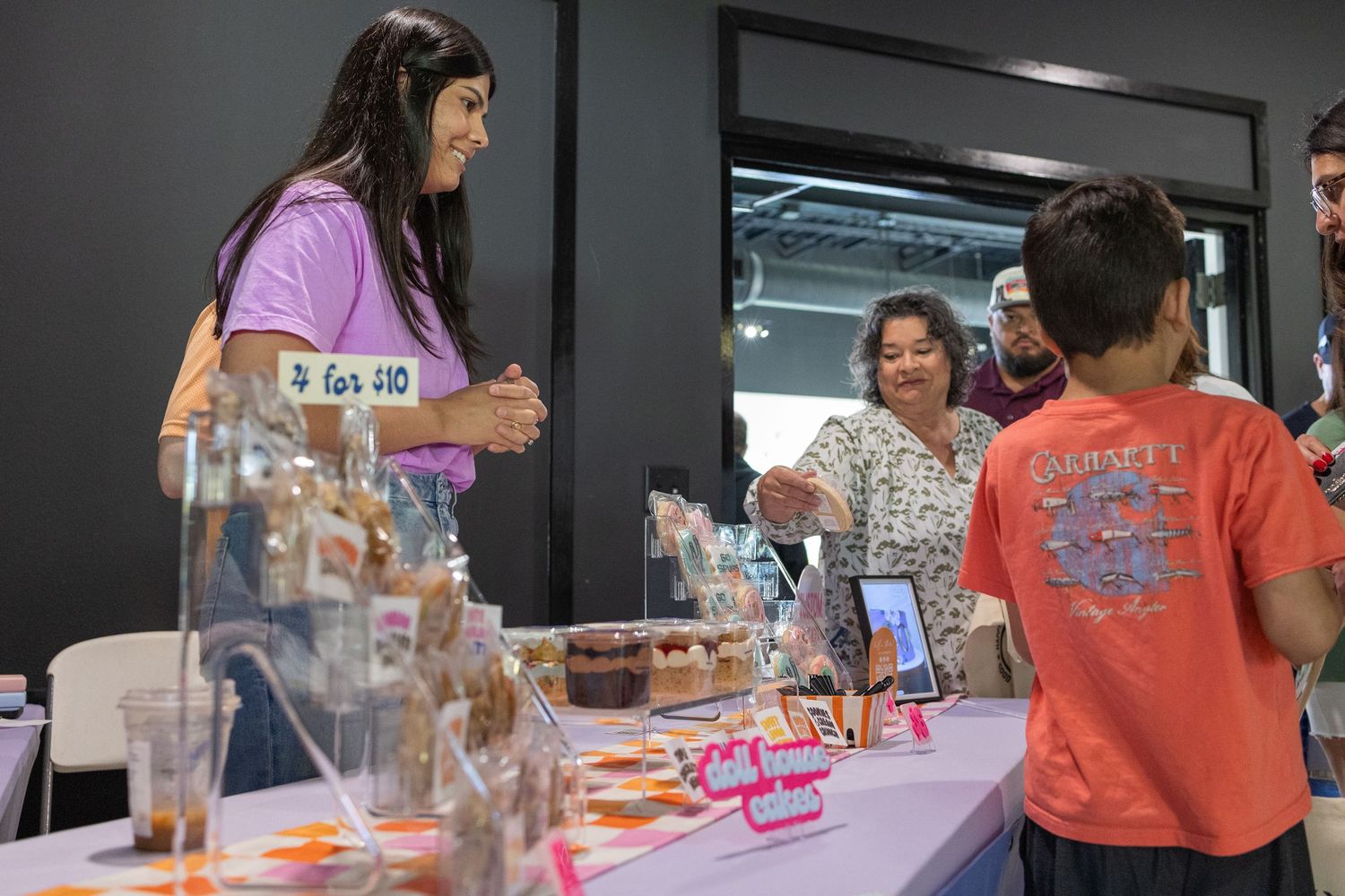 Woman vendor at a market table displays goods to a woman and child at an indoor event.
