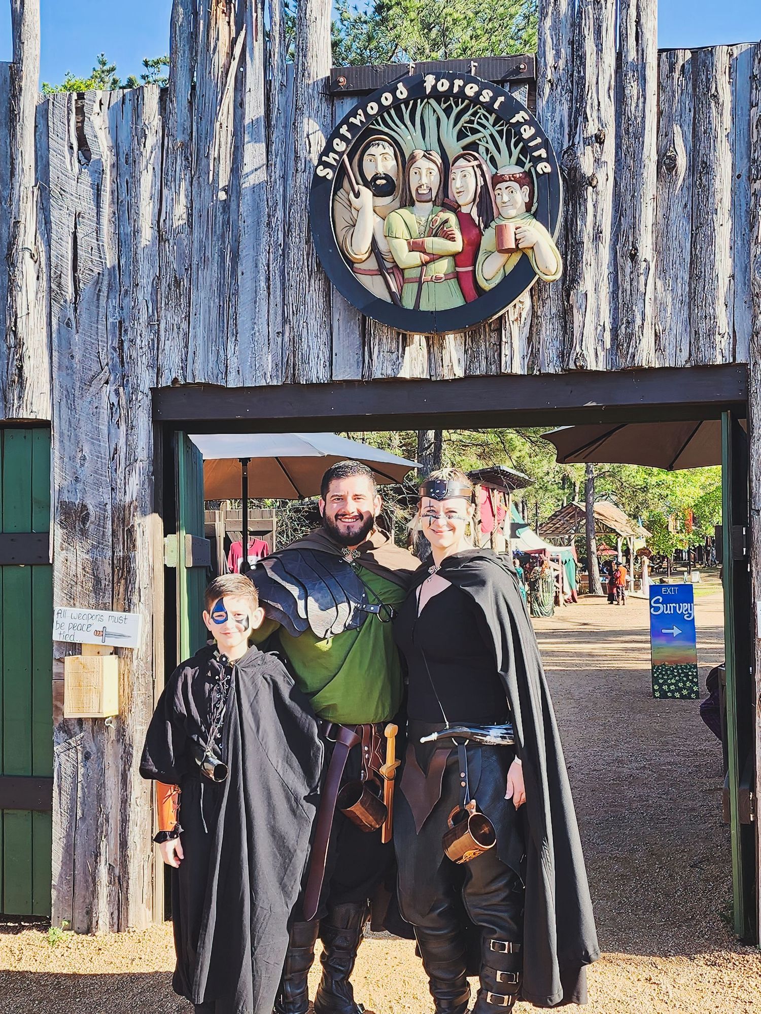 People in medieval costumes and cloaks gather at a rustic wooden fence during a Renaissance festival.