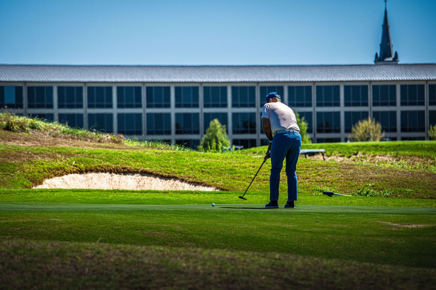 Golfer putting on a lush green golf course with a sand bunker and modern building in the background.