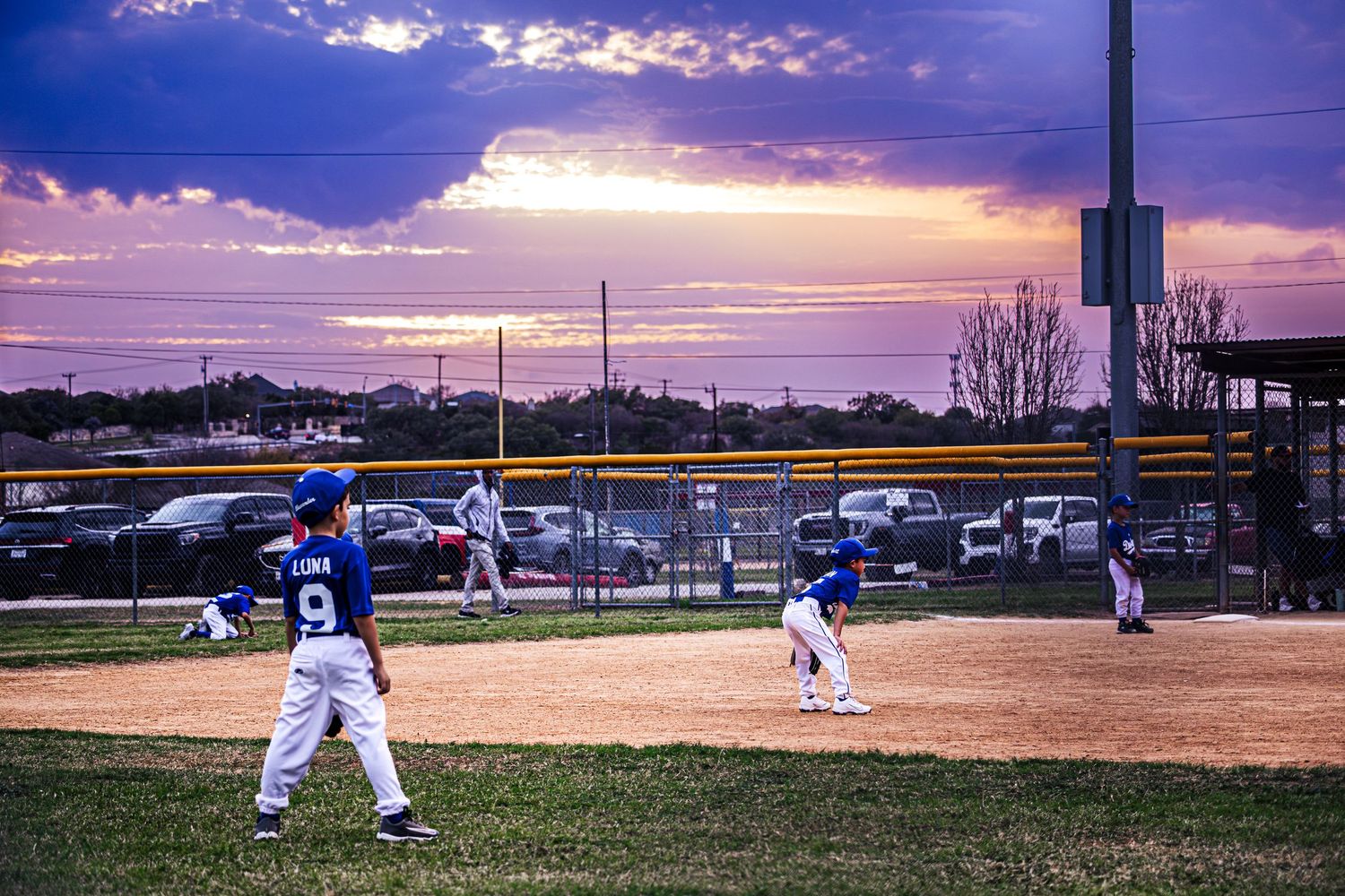 Youth baseball players in blue uniforms play a game at dusk under a dramatic purple and orange sunset sky.