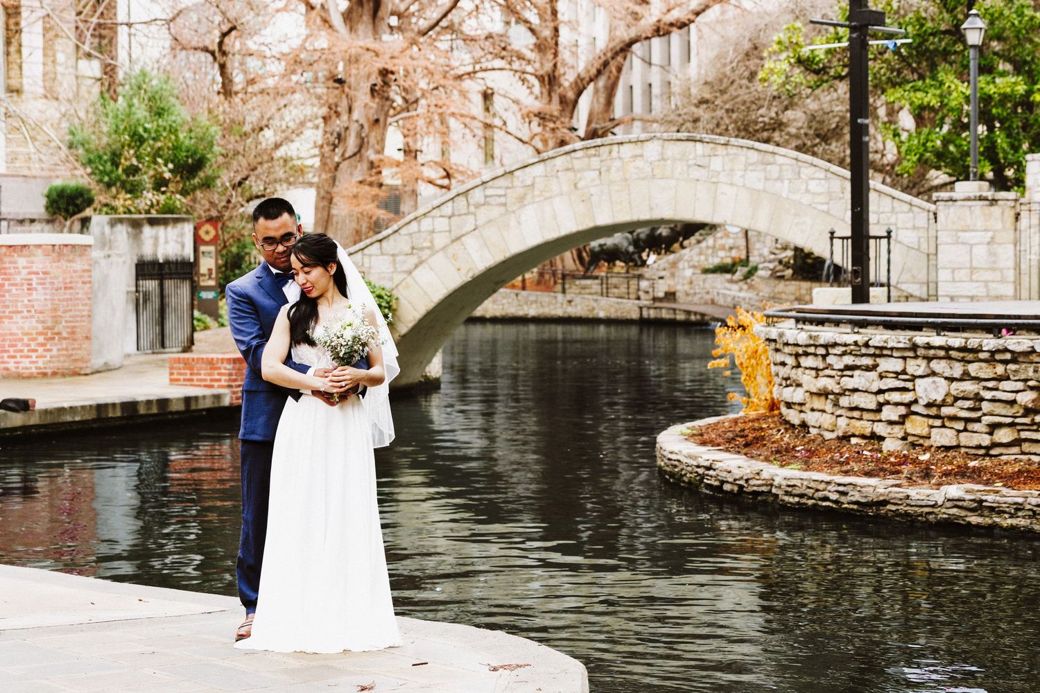 A couple in formal attire stands on a stone bridge over a peaceful canal in a romantic outdoor setting.