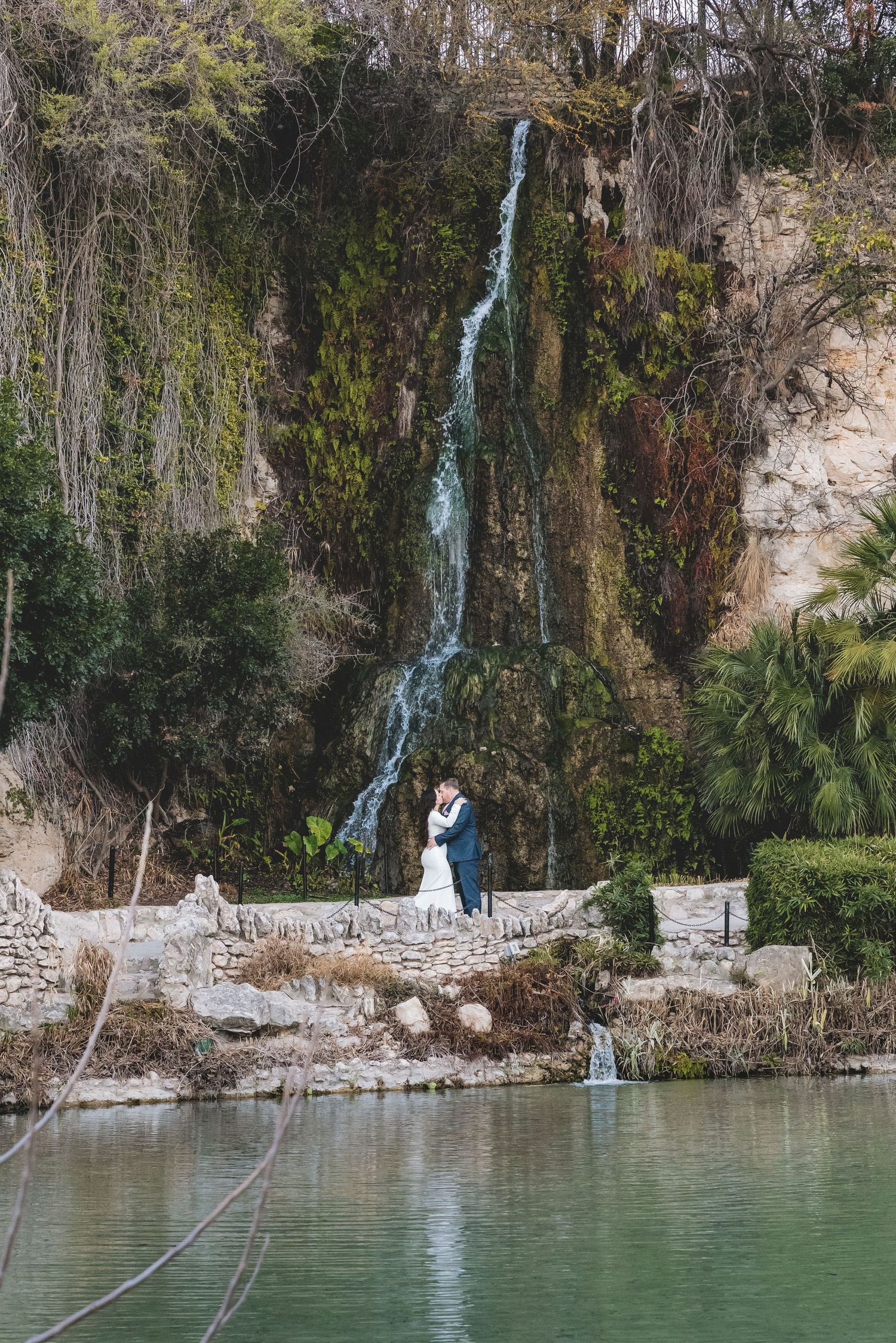 A waterfall cascades down a rocky cliff into a turquoise lake surrounded by lush vegetation and stone walls.