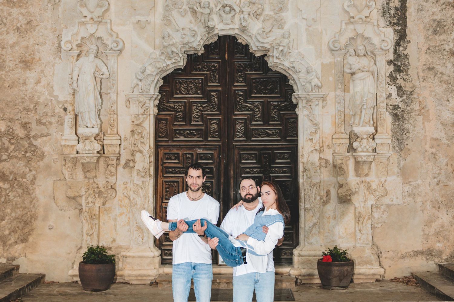 Family of three in white outfits holding babies before ornate wooden door at historic San Antonio Mission.