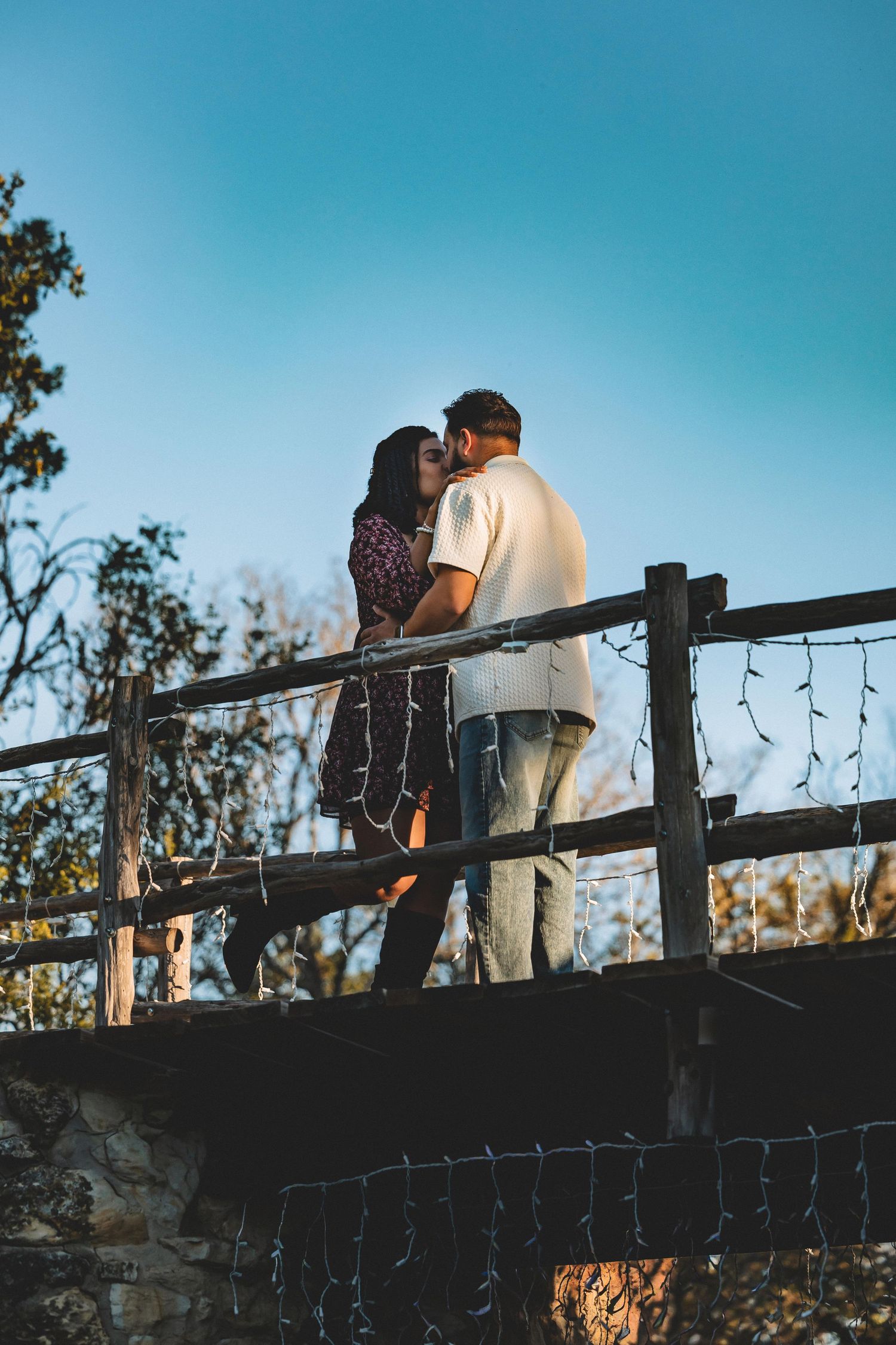 Silhouettes of a couple share an intimate moment against a bright blue sky on a wooden fence overlook.