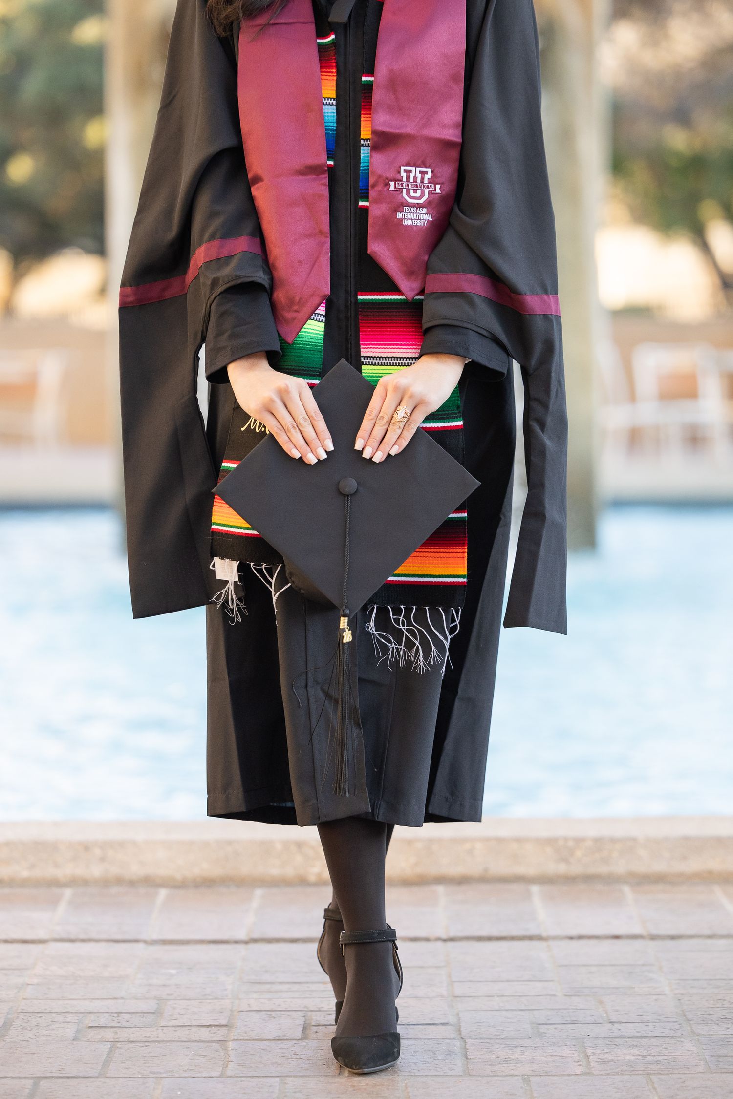 Graduate wearing black gown and colorful stole holds diploma during graduation ceremony by waterfront.