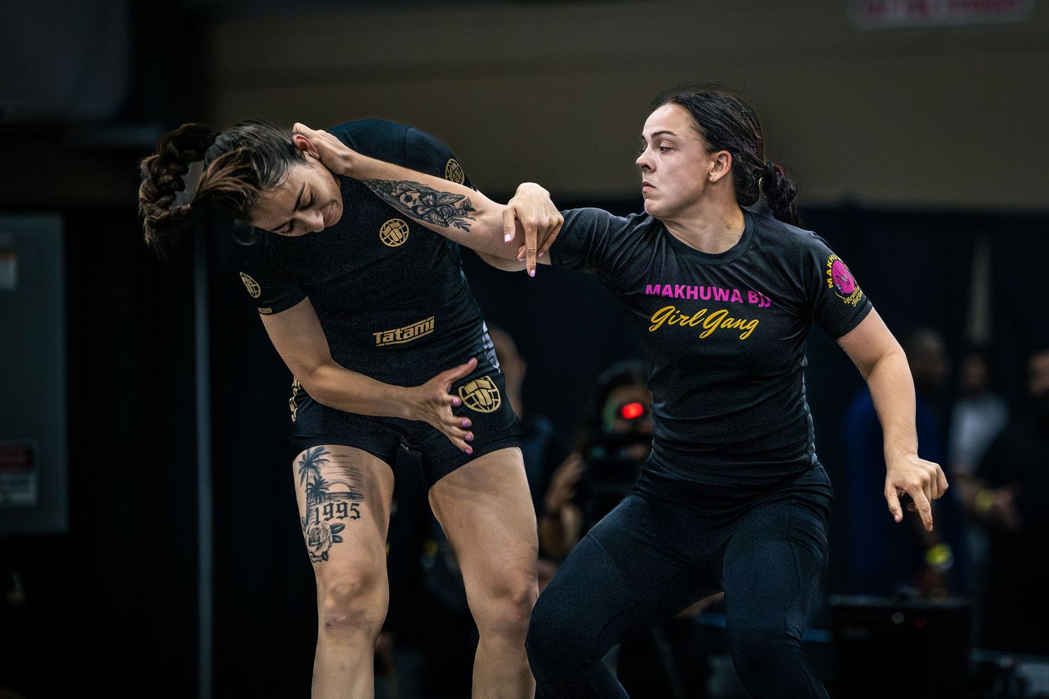 Two women compete in a grappling martial arts match, one wearing a Girl Gang shirt, in an indoor arena.