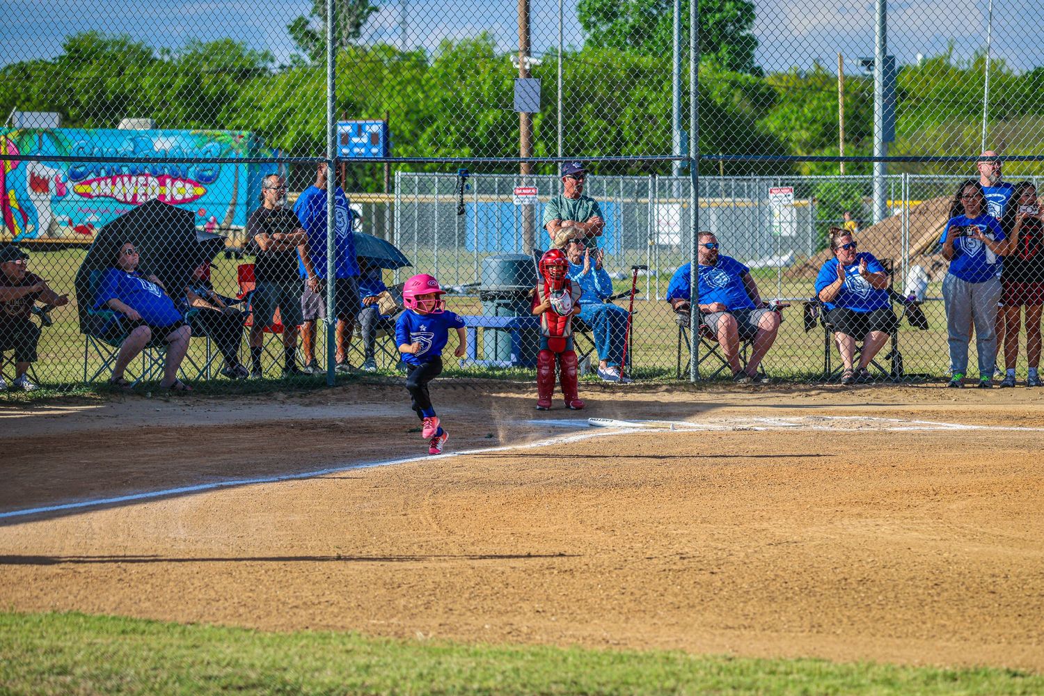 Young girl in pink helmet runs bases during a youth softball game with spectators watching from the dugout.