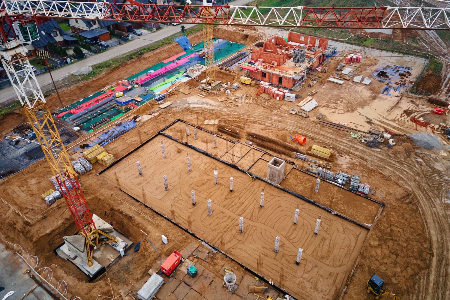 Aerial view of an active construction site with crane, foundation work, and building framework on red clay soil.