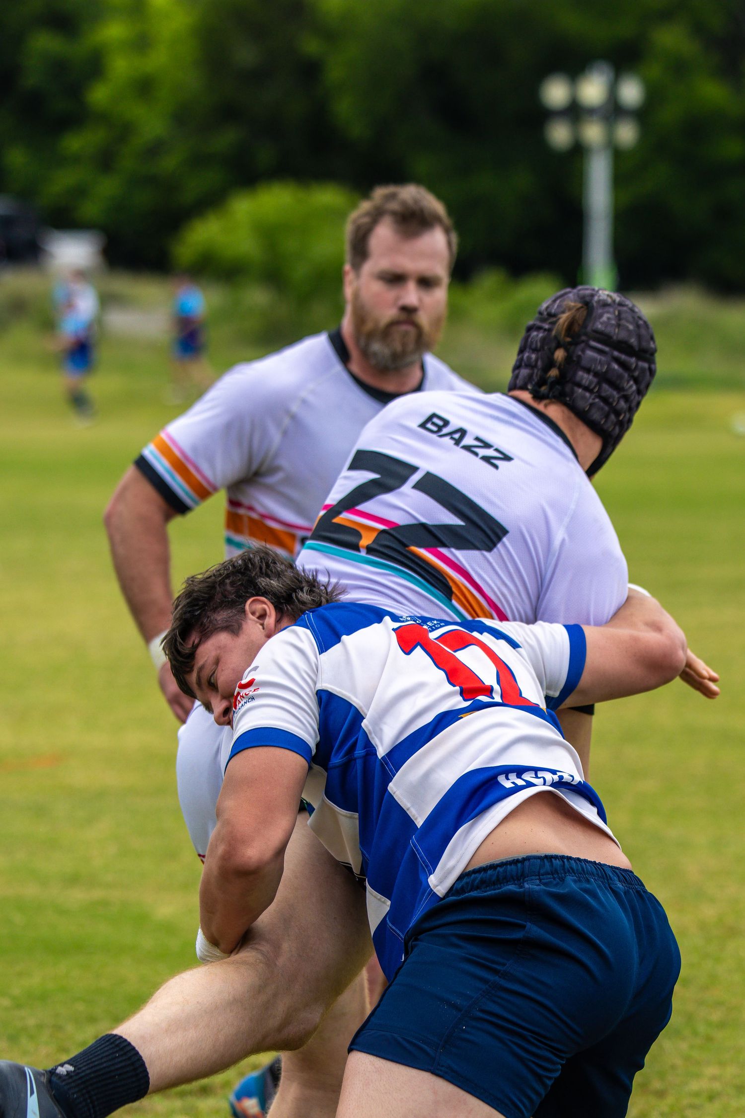 Rugby players in a intense tackle during a match, with a bearded player in white BAZZ jersey watching from behind.