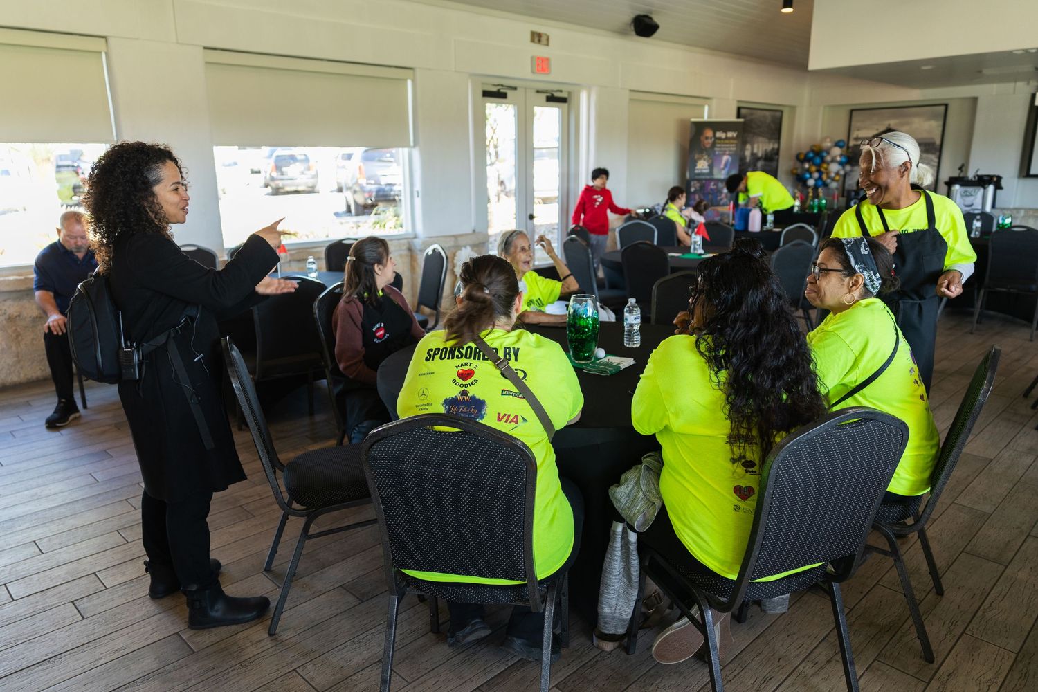 Group of people in neon yellow shirts seated at tables during an indoor meeting or training session.