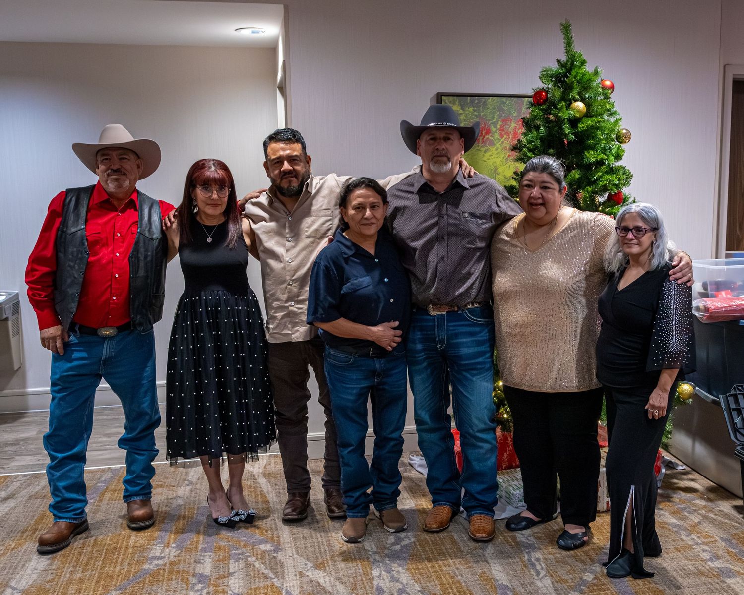 Seven people pose together smiling near a decorated Christmas tree in a festive indoor holiday gathering.