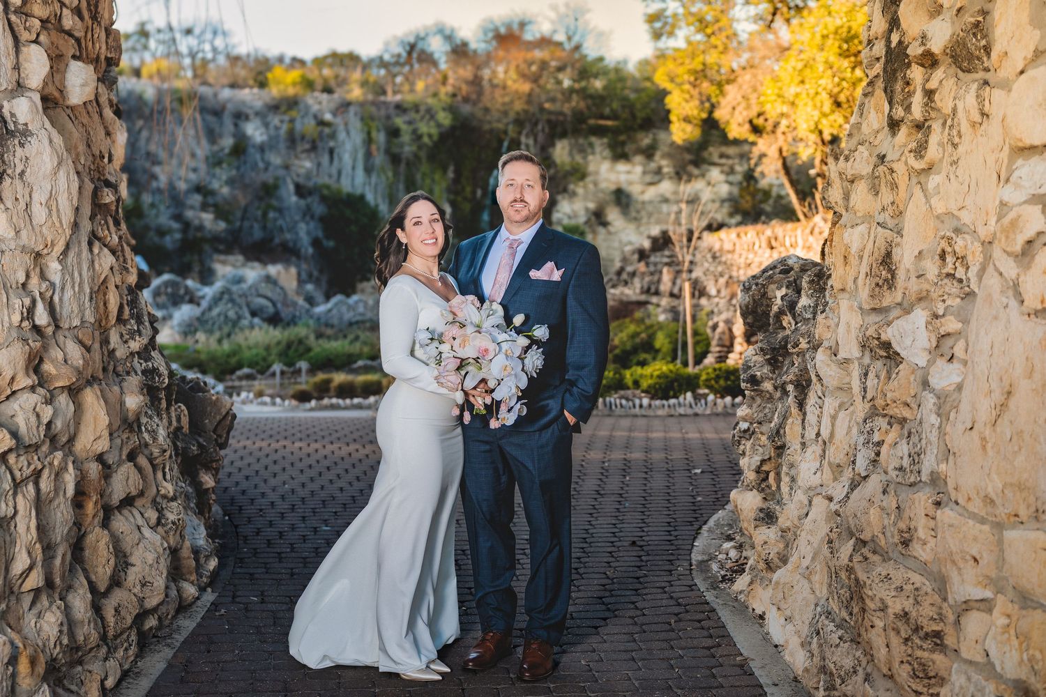 A couple in wedding attire poses between stone walls at an outdoor venue with autumn foliage in the background.