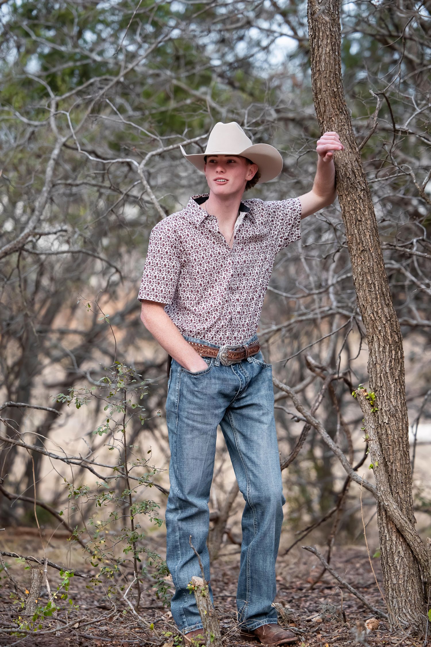 A cowboy in a patterned shirt and jeans poses casually against a tree in a wooded outdoor setting.