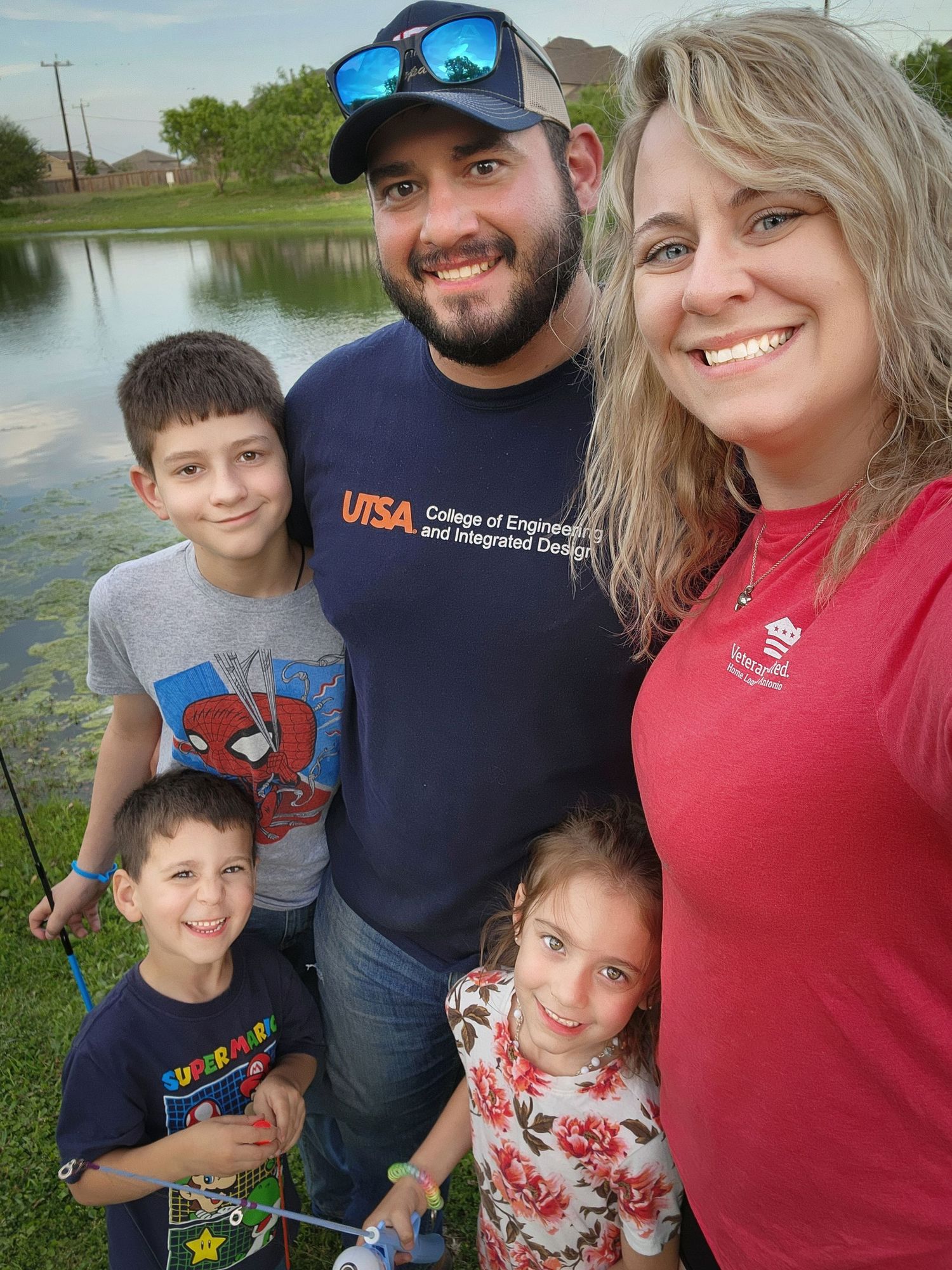A family takes a selfie together by a pond on a sunny day wearing casual clothes.