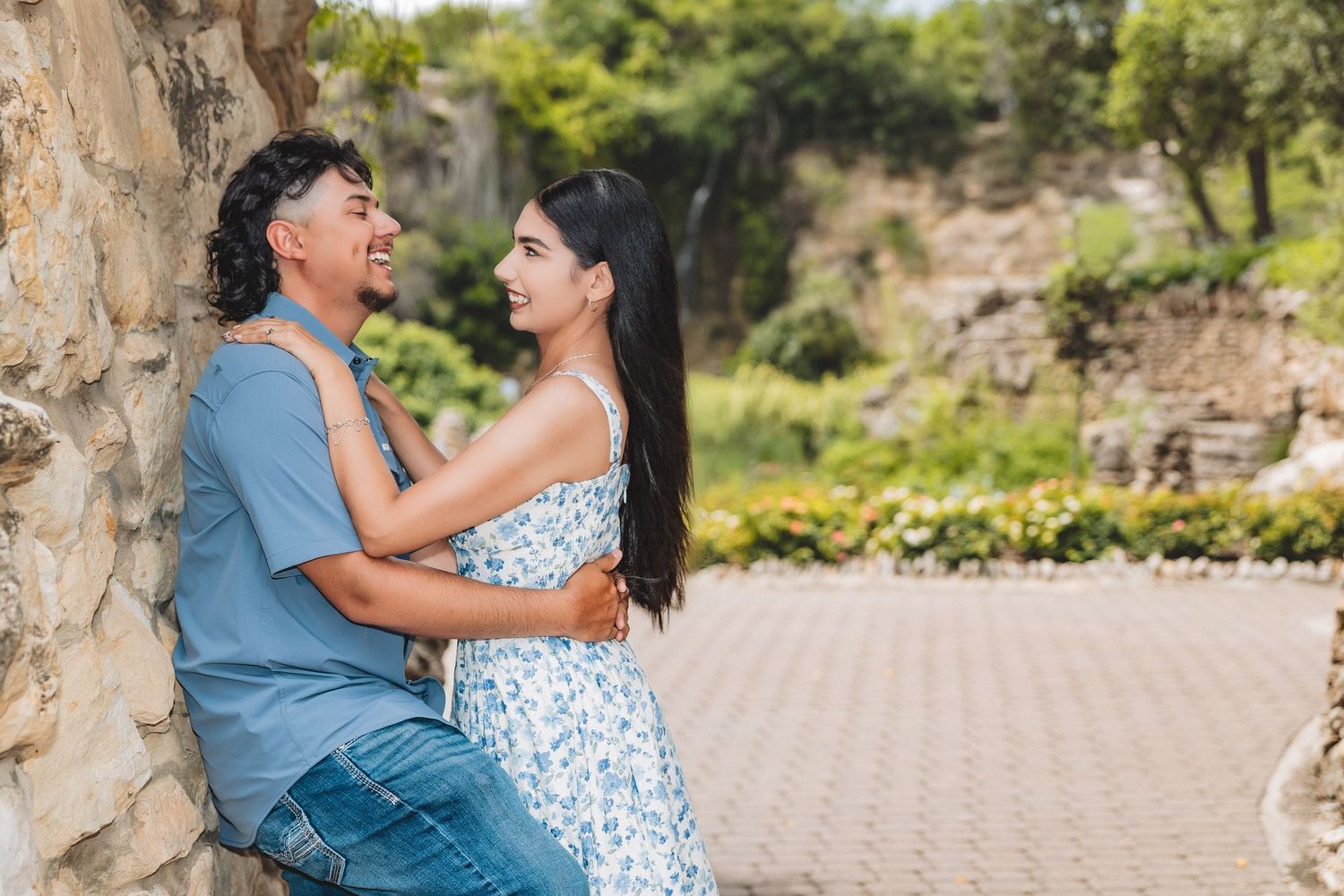 A young couple shares a romantic moment leaning against a stone wall in a lush outdoor garden setting.
