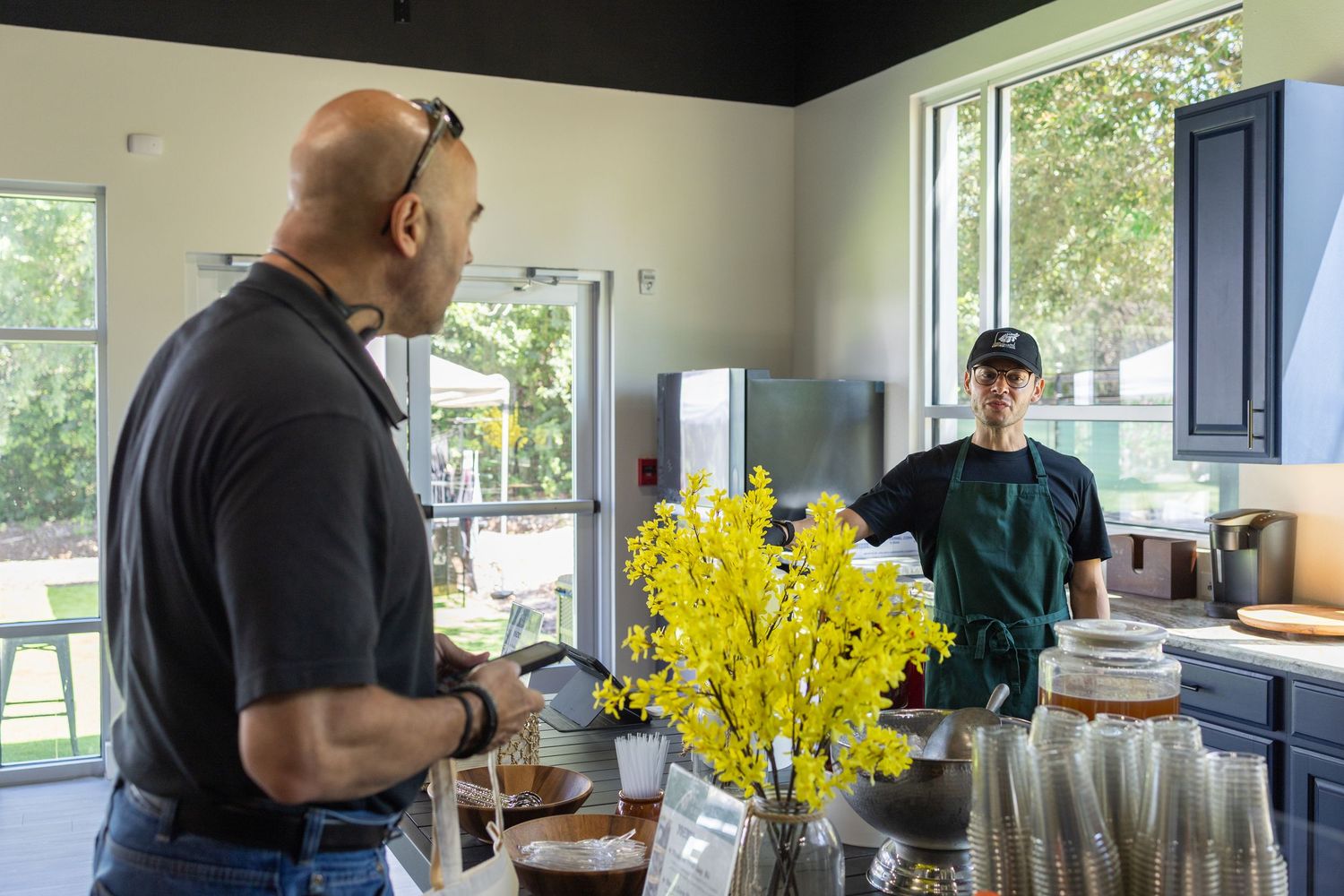 Bald man ordering from staff member at modern kitchen counter with yellow flowers.