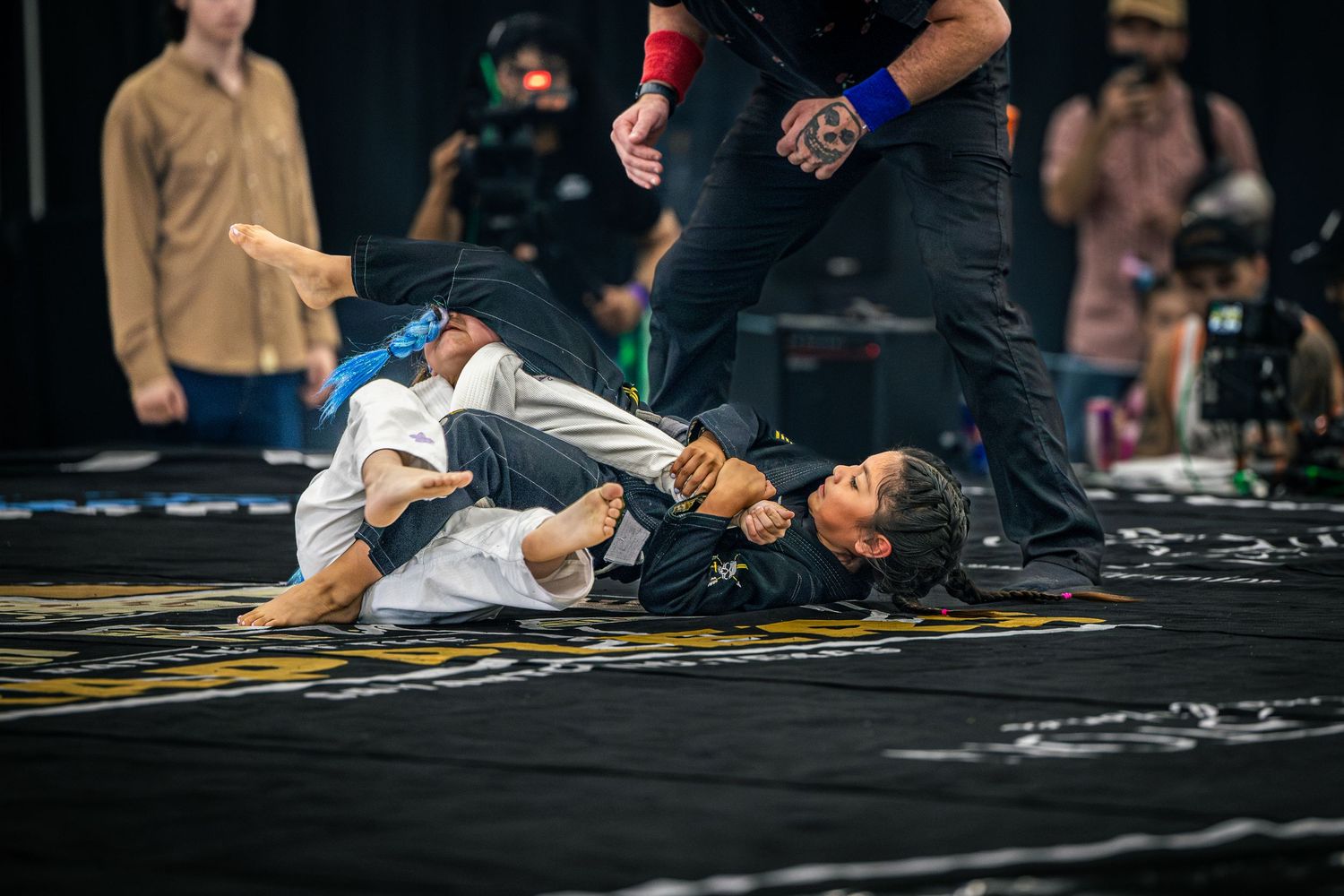 Two children competing in a Brazilian jiu-jitsu grappling match on a black mat at an indoor tournament.