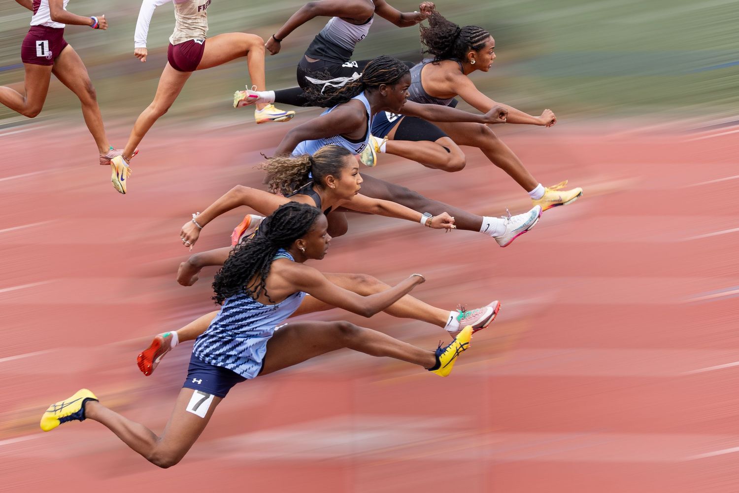 Female athletes sprint on a red track, captured in motion blur during a competitive race.