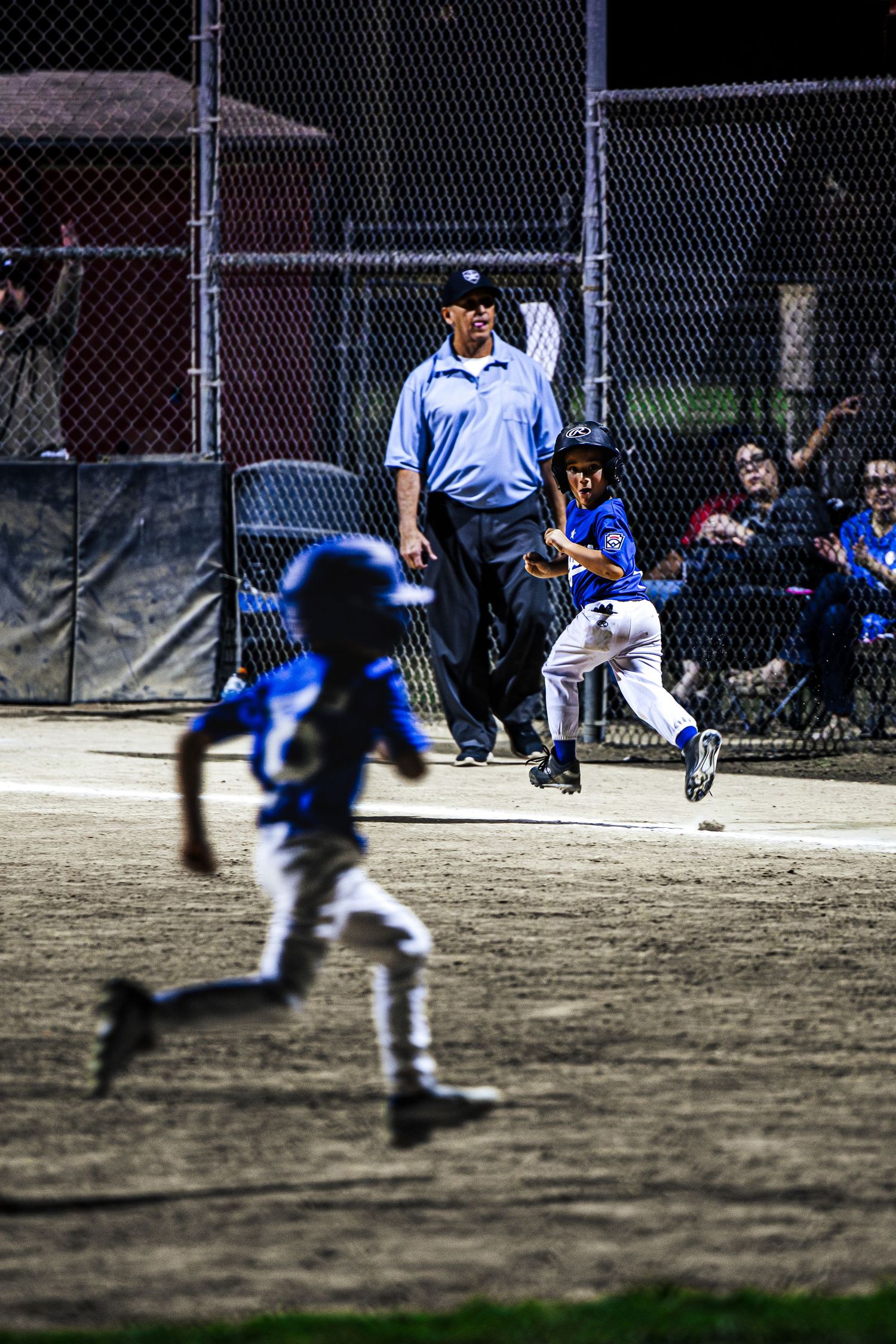 Young baseball players in blue uniforms compete in a night little league game as an umpire watches closely.