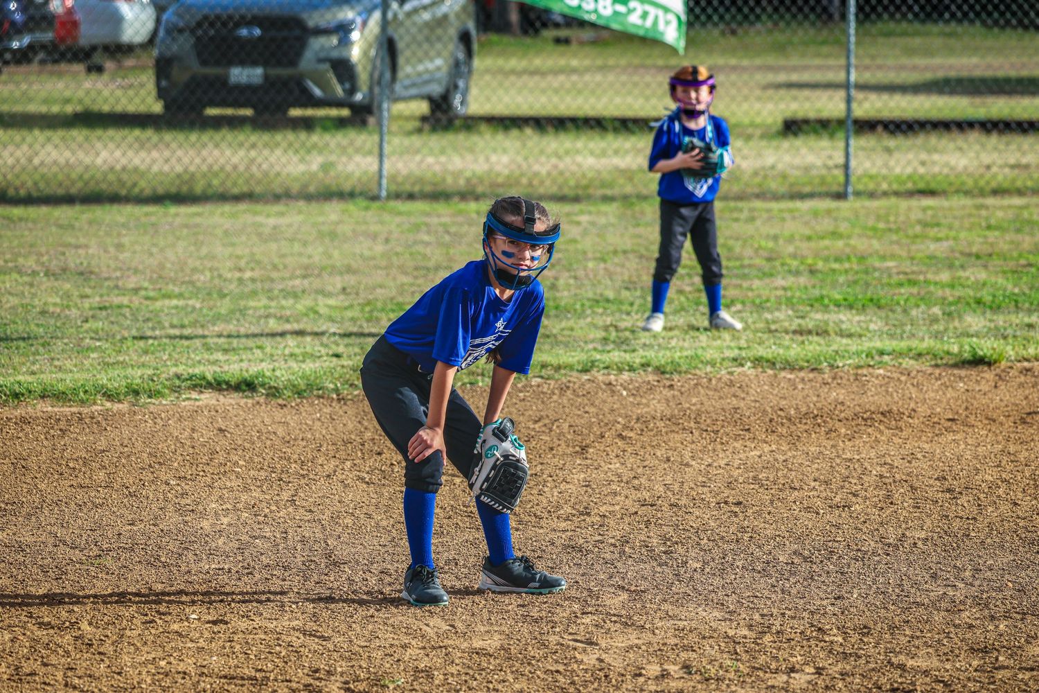 Young baseball player in blue uniform crouches in ready position on dirt infield during youth league game.
